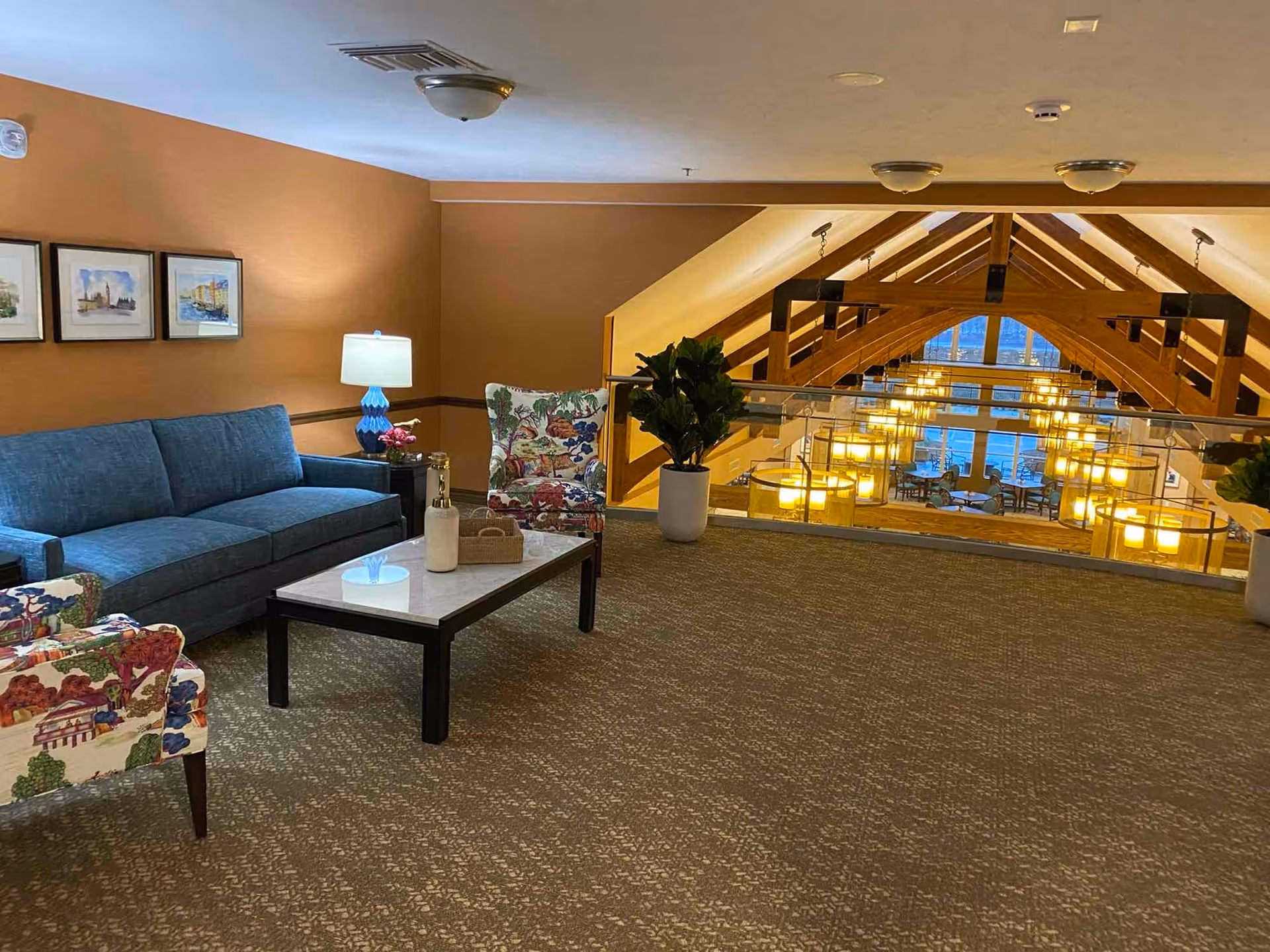 A cozy seating area with a blue sofa, two floral armchairs, a coffee table with decorative items, and a blue lamp on a side table. The space overlooks a large room with wooden beams and multiple hanging lights, visible through a glass railing.