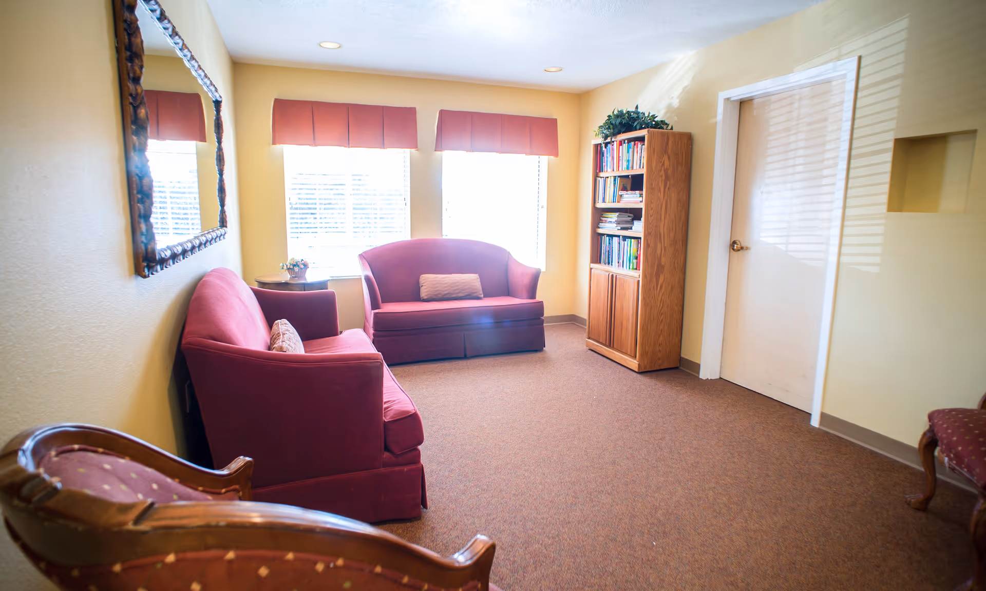 A cozy sitting room with two red upholstered loveseats, a wooden bookshelf filled with books, a small side table with a flower arrangement, a large decorative mirror on the wall, and two windows with red valances letting in natural light.