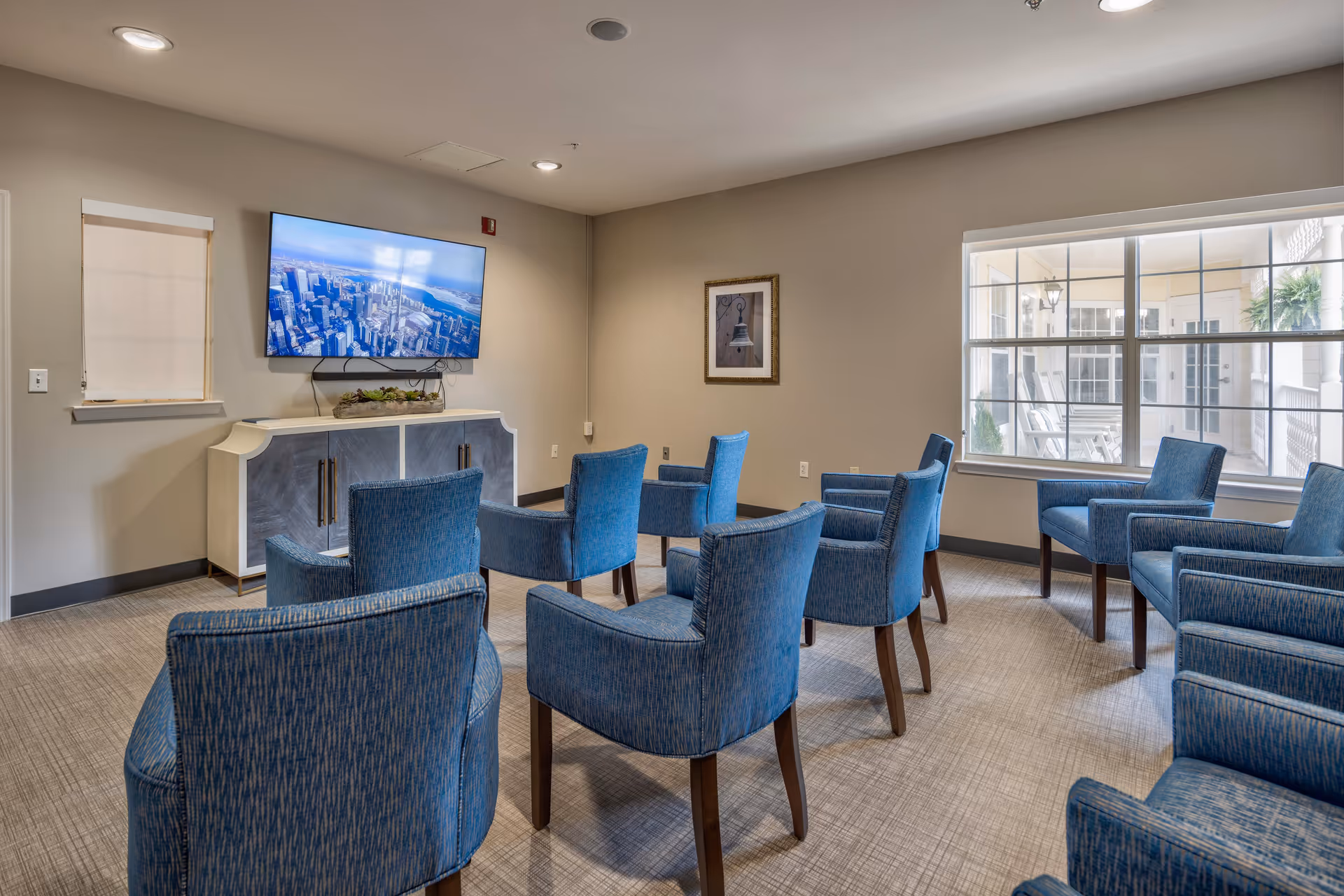 A small meeting or media room with multiple blue upholstered chairs arranged in rows facing a wall-mounted flat screen TV. The room has beige walls, a large window showing an outdoor patio with rocking chairs, and a cabinet beneath the TV with decorative plants on top.