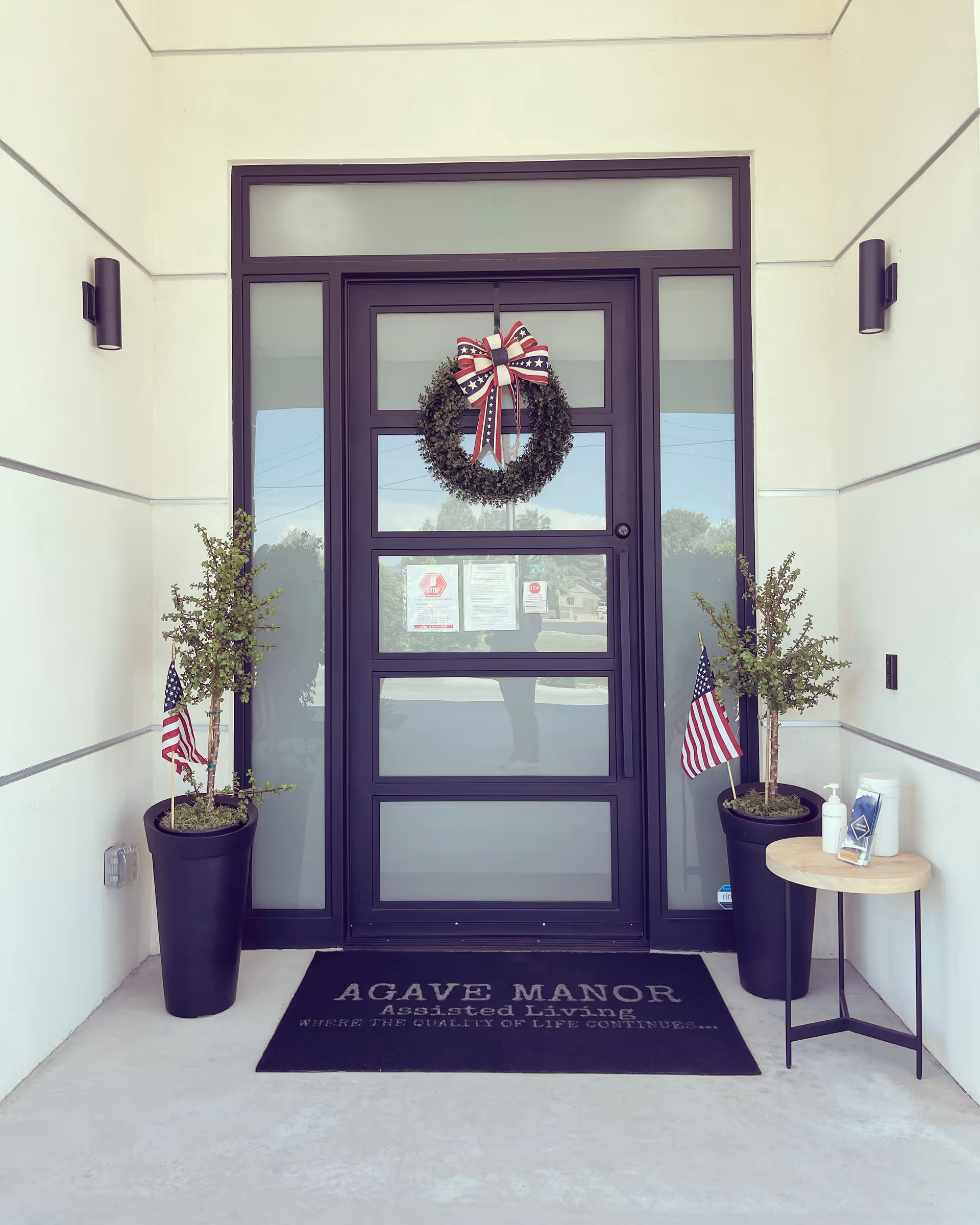 Entrance door of Agave Manor Assisted Living facility with a patriotic wreath hanging on the door. Two potted plants with small American flags flank the door. A small round table on the right holds hand sanitizer and wipes. A black doormat with the text 'AGAVE MANOR Assisted Living WHERE THE QUALITY OF LIFE CONTINUES...' is placed in front of the door.