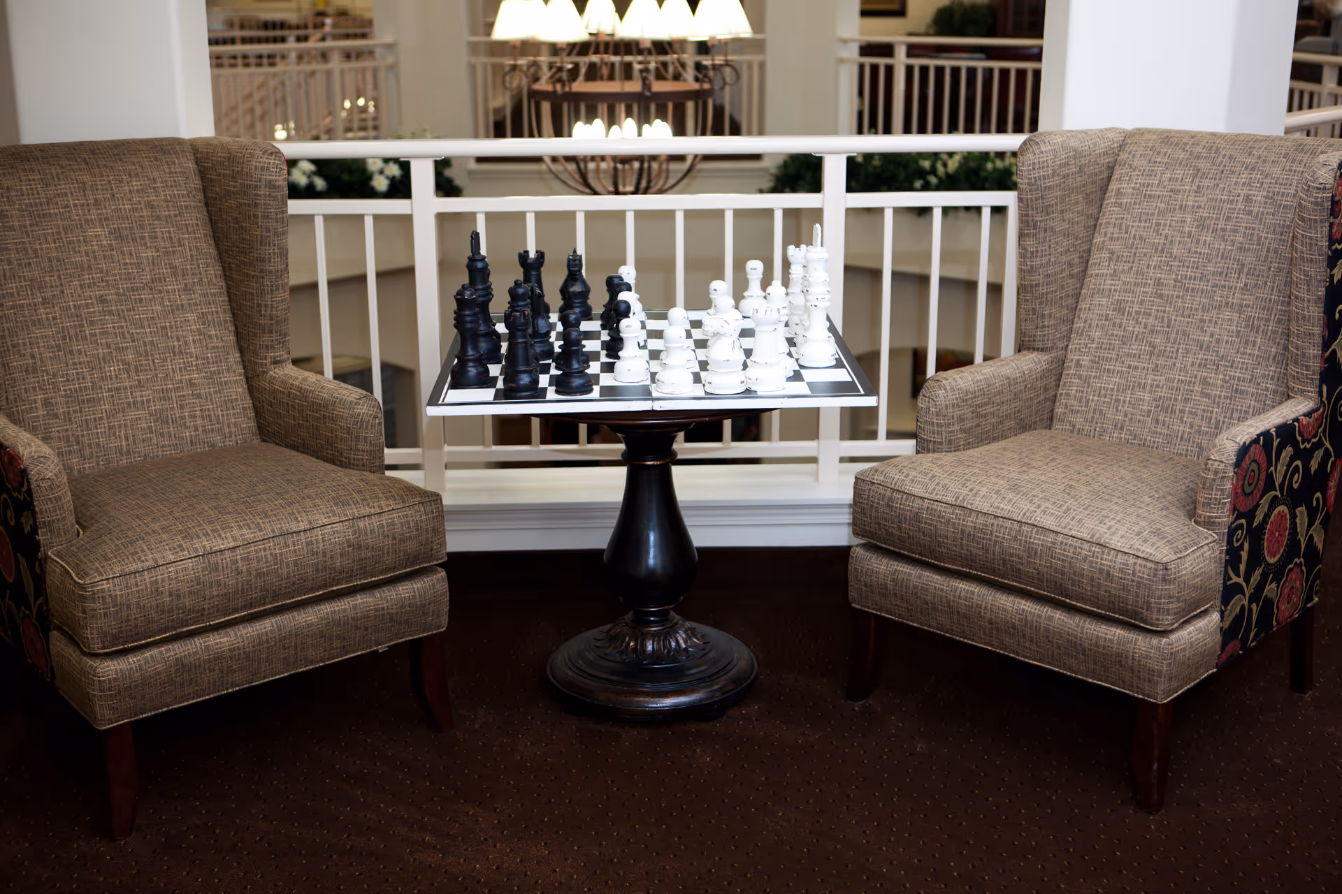 Two upholstered armchairs face a small pedestal table with a chessboard set between them in an indoor seating area.