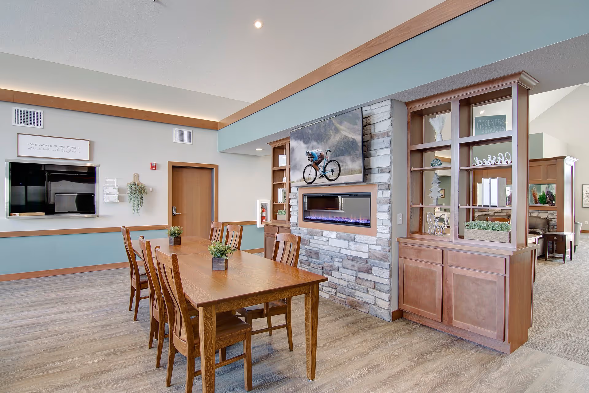 A dining area with a wooden table and six chairs, decorated with small potted plants. The room features a stone fireplace with a modern electric insert and a mounted flat-screen TV above it displaying a cyclist. There are built-in wooden shelves and cabinets on either side of the fireplace. The walls are painted light gray with a teal accent below the chair rail, and the floor is wood laminate. A door and a wall-mounted TV are visible in the background.