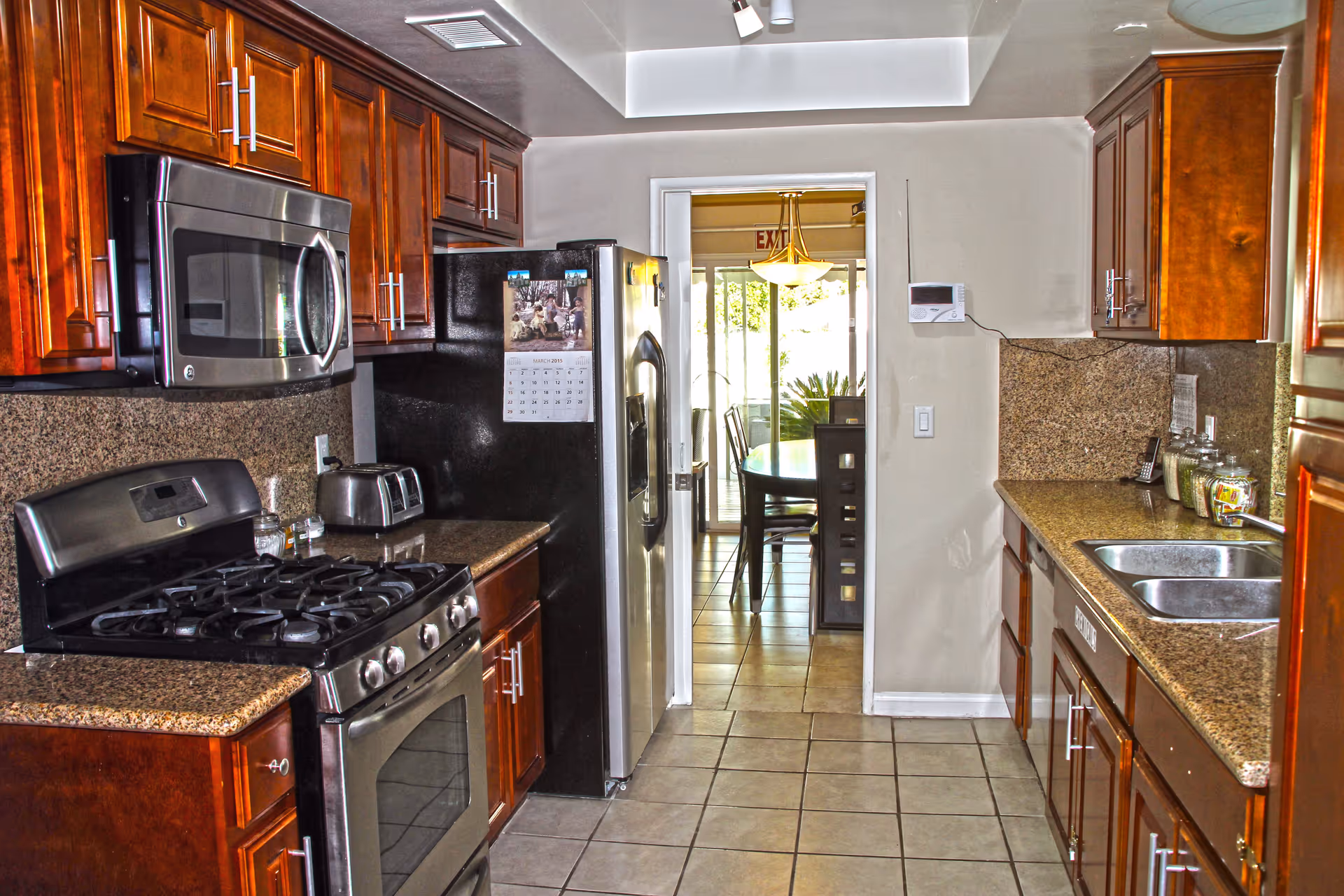 A kitchen with wooden cabinets, granite countertops, a stainless steel gas stove with oven, a microwave, a black refrigerator with a calendar on the door, a toaster, and a double sink. The floor is tiled, and there is a doorway leading to a dining area with a table and chairs visible.