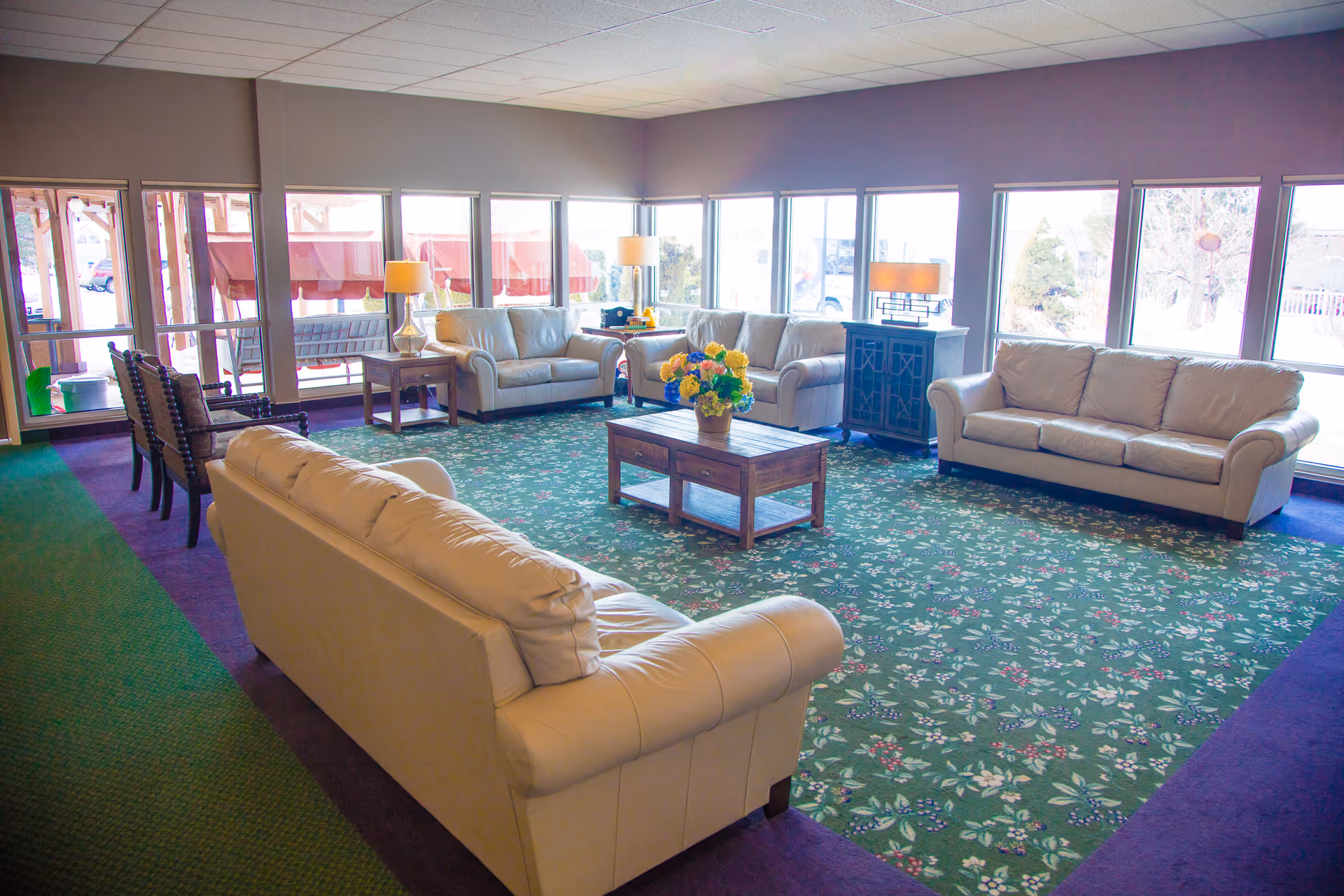 A bright communal lounge with beige leather sofas arranged around a wooden coffee table on a patterned green carpet and large windows.