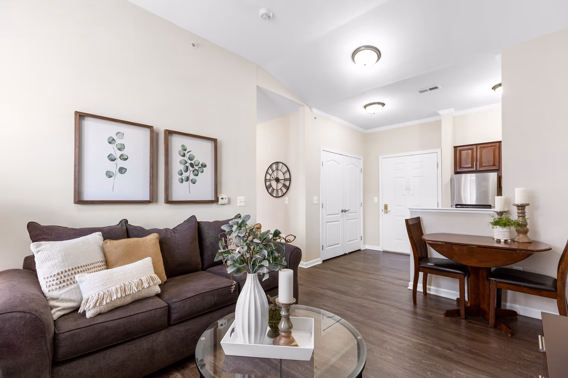 A cozy living area with a dark brown sofa adorned with beige and white pillows. Above the sofa are two framed botanical prints. In front of the sofa is a round glass coffee table with a white tray holding a white vase with green foliage and a candle. To the right, there is a small wooden dining table with two chairs, decorated with candles and a small plant. The background shows a kitchen area with wooden cabinets and a stainless steel refrigerator, and a white door leading to another room. The floor is dark wood, and the walls are painted light beige.