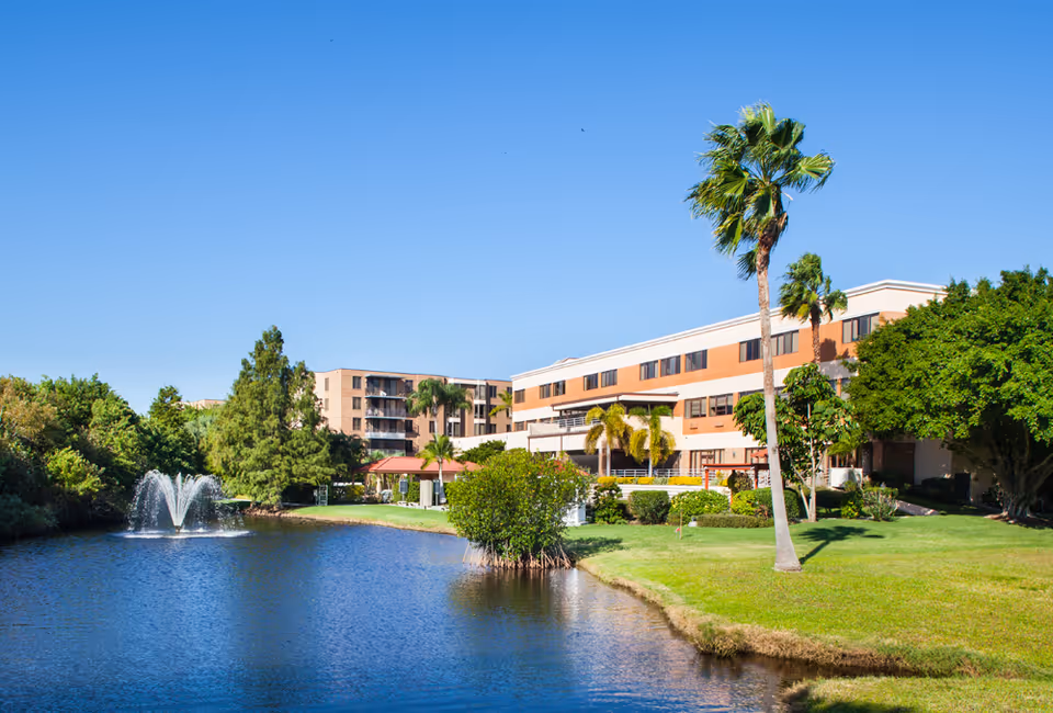 A multi-story building sits beside a pond with a fountain, surrounded by palm trees and a manicured lawn under a clear blue sky.