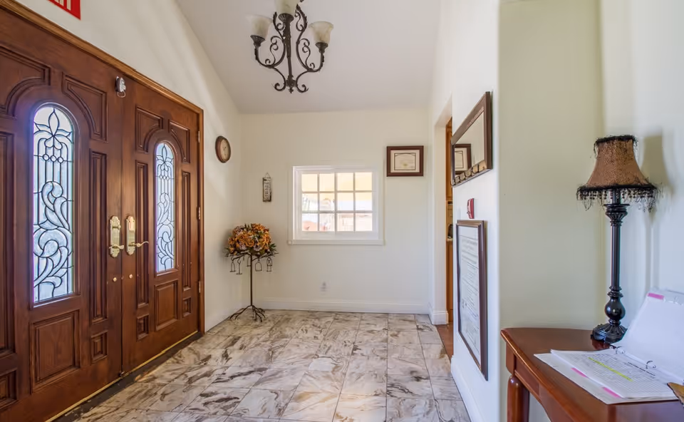 Entrance area with double wooden doors featuring decorative glass panels, a tiled floor, a small window on the far wall, a floral arrangement on a stand, a chandelier hanging from the ceiling, and a wooden table with a lamp and open binder on the right side.