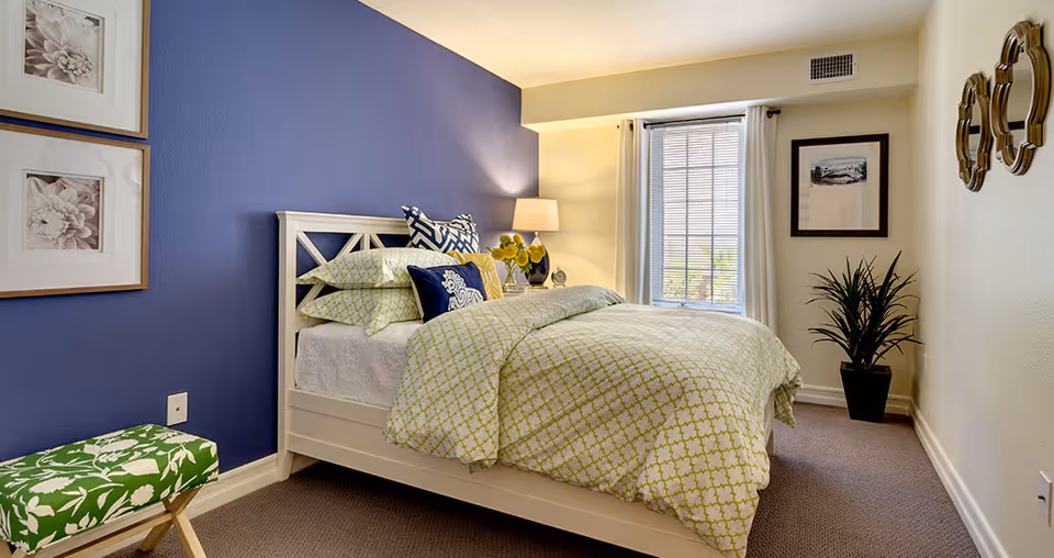A cozy bedroom with a white bed frame and green patterned bedding. The room features a blue accent wall with two framed floral pictures, a green patterned bench, a bedside table with a lamp and flowers, a window with white curtains, a potted plant, and decorative mirrors on the opposite wall.