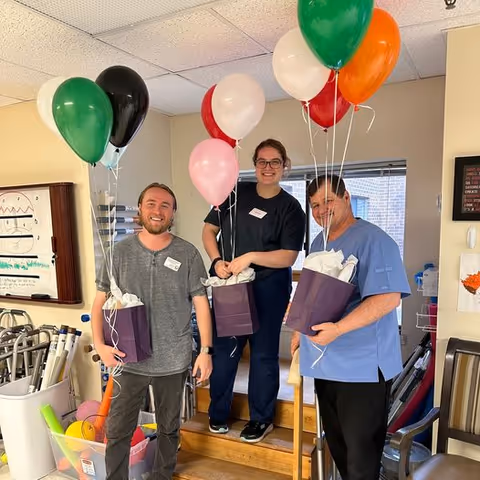 Three smiling people standing indoors holding purple gift bags with colorful balloons attached. The setting appears to be a nursing or care facility with medical and recreational equipment visible in the background.