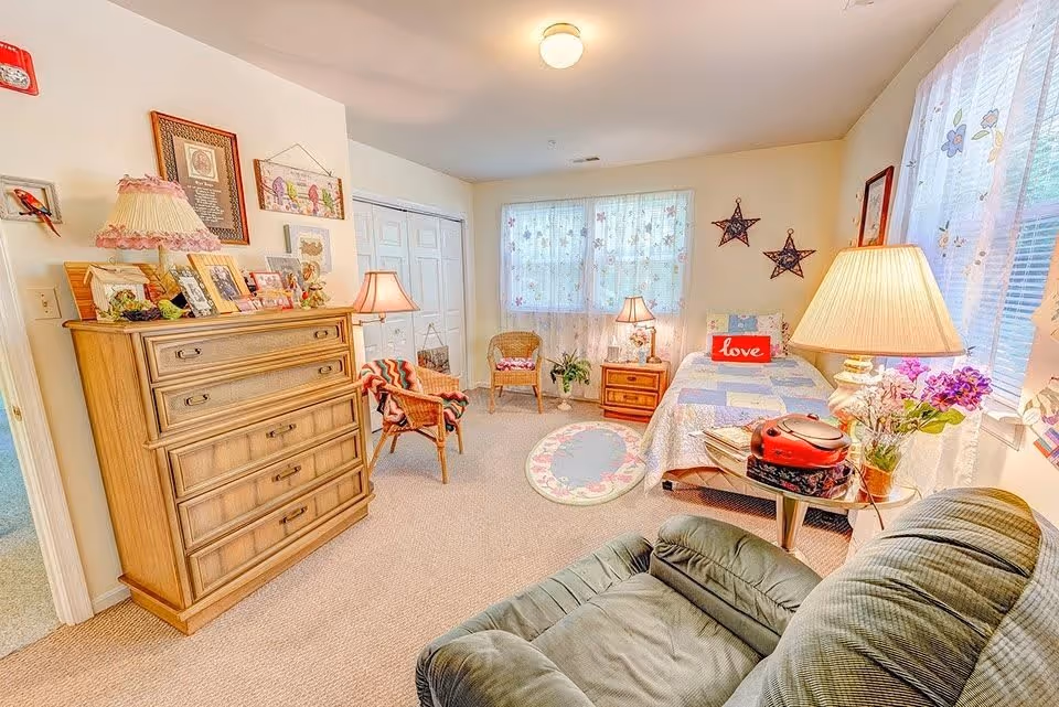 A cozy bedroom with a single bed covered in a patchwork quilt and a red pillow with the word 'love'. The room has two wicker chairs, a wooden dresser with framed photos and decorative items, two table lamps, a small round rug, and a green recliner chair. The windows are adorned with sheer floral curtains, and the walls have framed pictures and star-shaped decorations.
