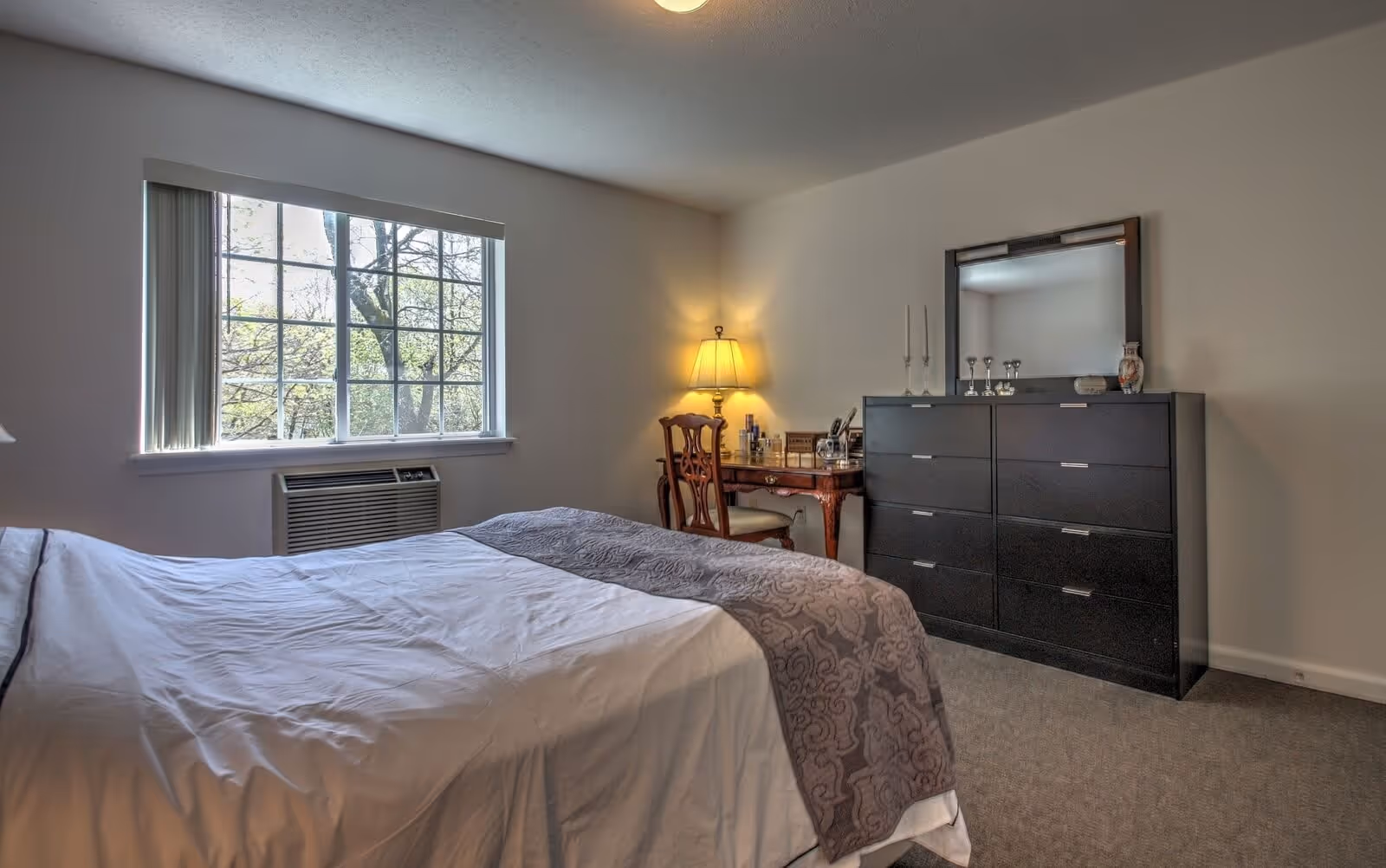 A bedroom with a large window showing trees outside, a bed with white and gray bedding, a wooden chair and desk with a lit table lamp, and a black dresser with a mirror and decorative items on top.