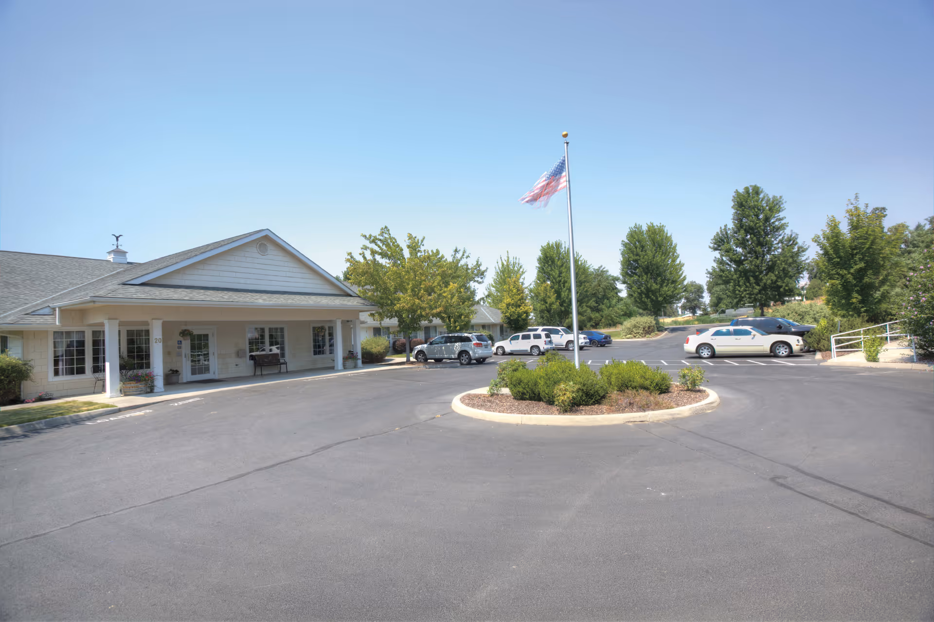Exterior view of a single-story building with a covered entrance and several windows. There is a circular driveway with a landscaped island in the center featuring bushes and an American flag on a flagpole. Several cars are parked in the parking lot surrounded by trees and greenery under a clear blue sky.