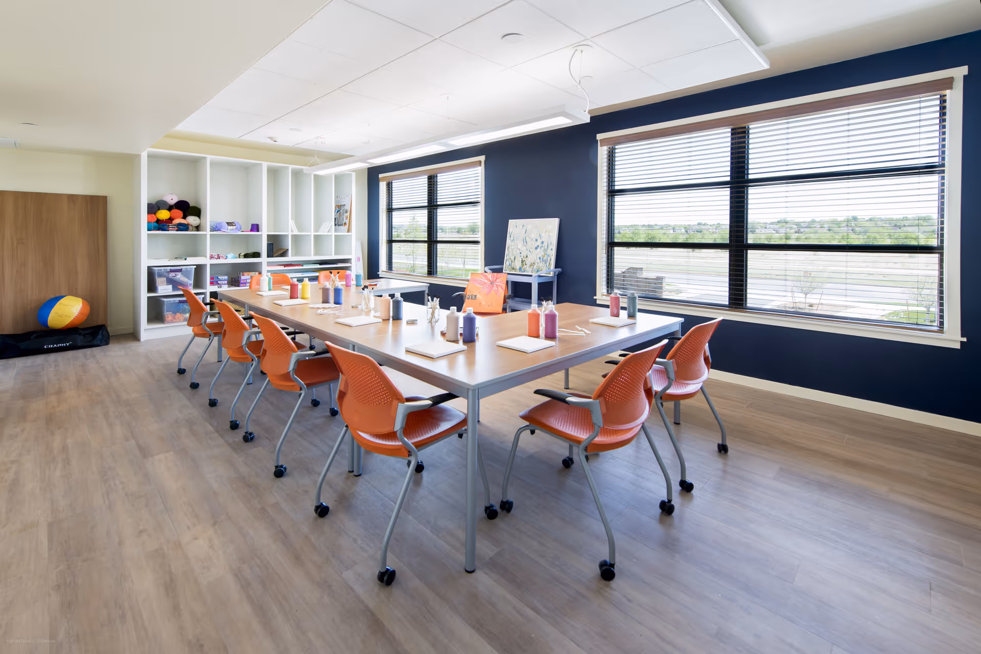 A bright, spacious activity room with a large rectangular table surrounded by orange chairs on wheels. The table is set with paint bottles, brushes, and canvases, suggesting an art or craft session. The room has large windows with blinds, letting in natural light, and white shelves filled with craft supplies and colorful yarn balls. The walls are painted dark blue and white, and the floor is light wood.