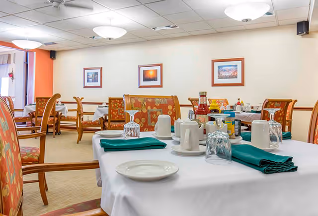 A dining room in Brenden Gardens facility with tables covered in white tablecloths, set with plates, upside-down glasses, mugs, green cloth napkins, and condiments. The room has wooden chairs with patterned upholstery, framed pictures on the wall, and ceiling lights.