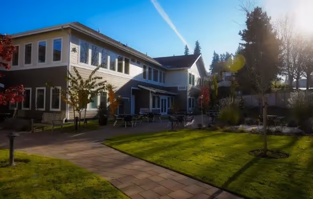 Outdoor view of a senior living facility with a two-story building, paved walkways, green lawn, trees, and outdoor seating under a clear blue sky.