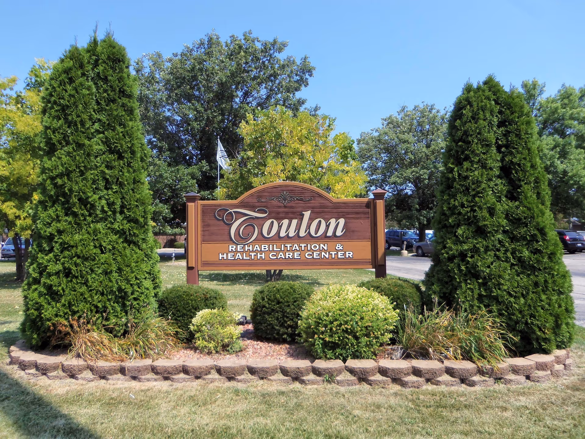 A wooden sign reading 'Toulon Rehabilitation & Health Care Center' surrounded by neatly trimmed bushes and tall evergreen trees, with a parking lot and cars visible in the background under a clear blue sky.