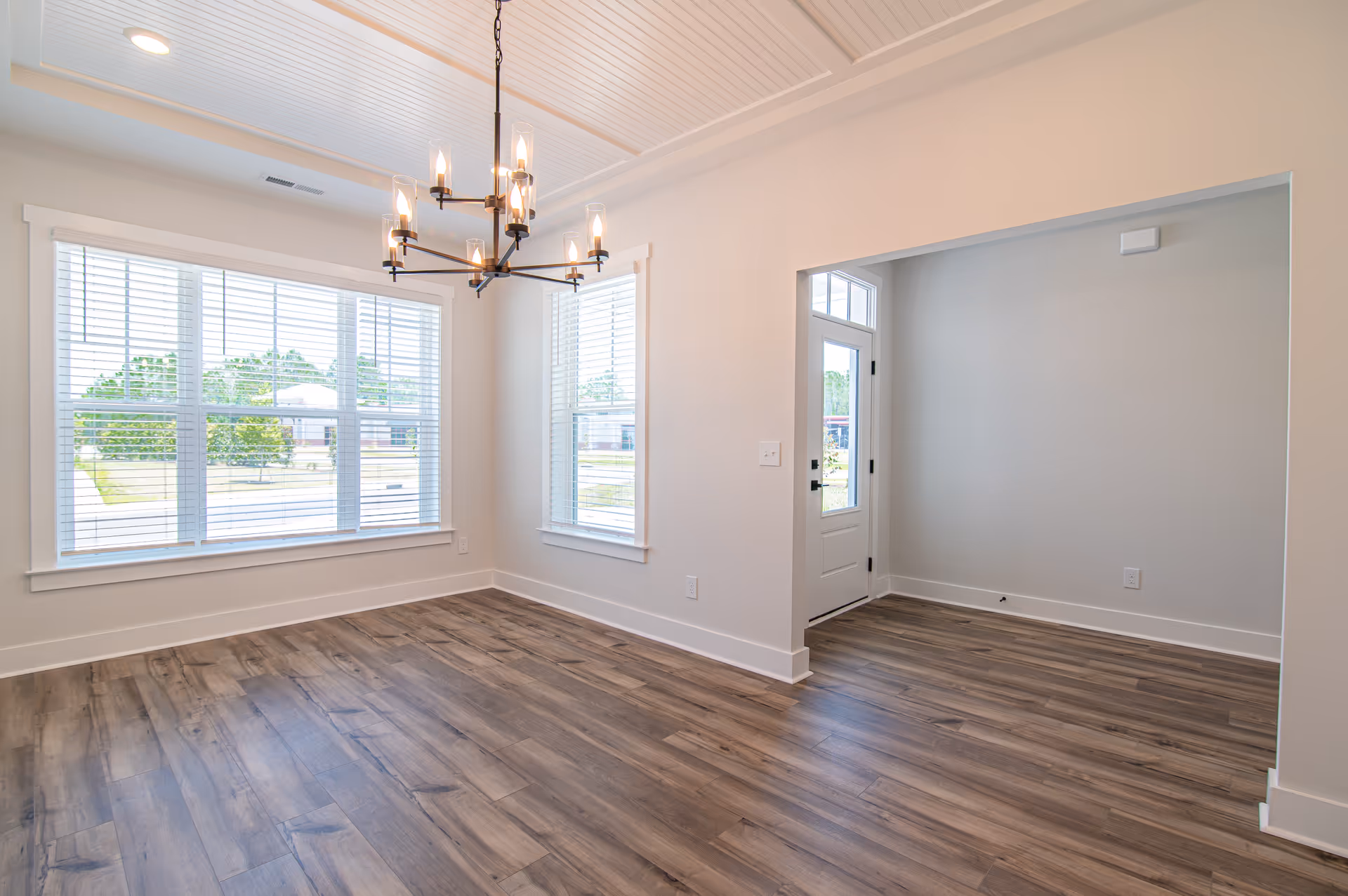 Bright empty dining room with wood floors, a chandelier, large windows with blinds, and an entry door.