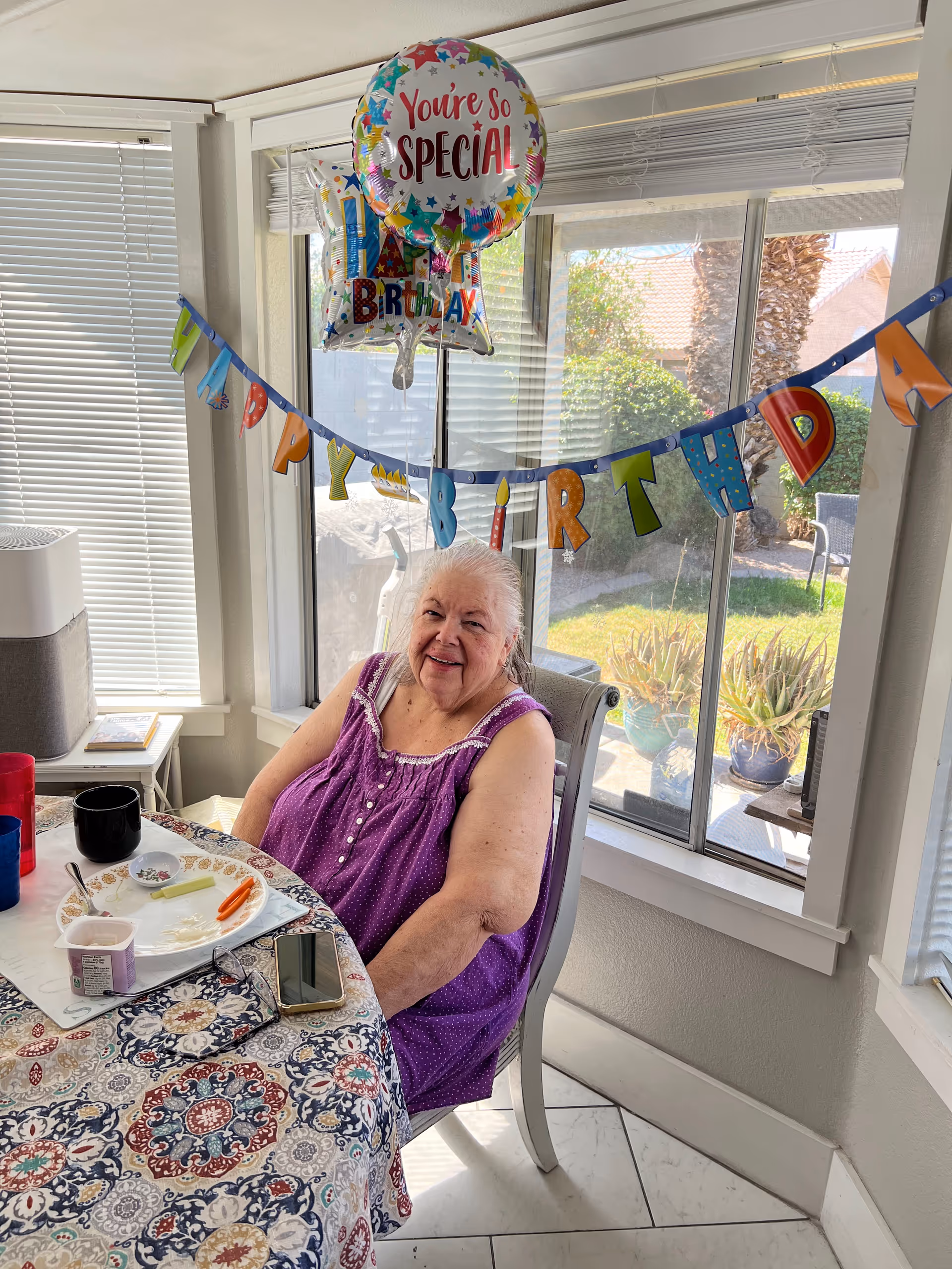 An elderly woman wearing a purple sleeveless top is sitting at a dining table with a colorful patterned tablecloth. Behind her, there are birthday decorations including a banner that says 'HAPPY BIRTHDAY' and balloons, one of which reads 'You're So Special'. The room has large windows showing a sunny outdoor garden with plants and a palm tree.
