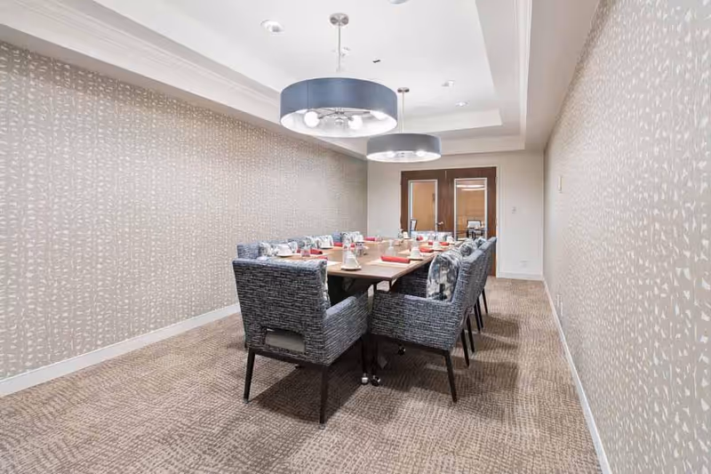 A modern dining room with a long rectangular table set for a meal, surrounded by cushioned chairs with patterned upholstery. The room features patterned wallpaper, carpeted floor, and two large circular pendant lights hanging from the ceiling. Double wooden doors are visible at the far end of the room.