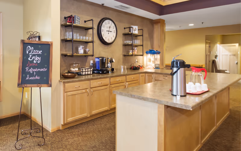Communal refreshment counter with coffee urns, a popcorn machine, shelves of glasses, and a chalkboard sign inviting guests to enjoy refreshments.