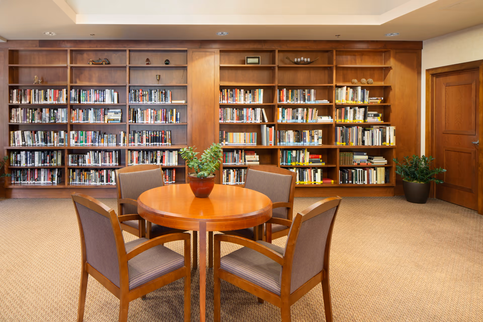 Wood-paneled library room with built-in bookshelves, a round wooden table with four chairs and a potted plant.