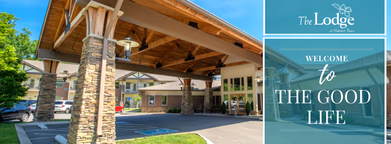 Entrance area of The Lodge at Natchez Trace featuring a covered drop-off with stone pillars and wooden beams, surrounded by parking spaces and residential-style buildings under a clear blue sky.