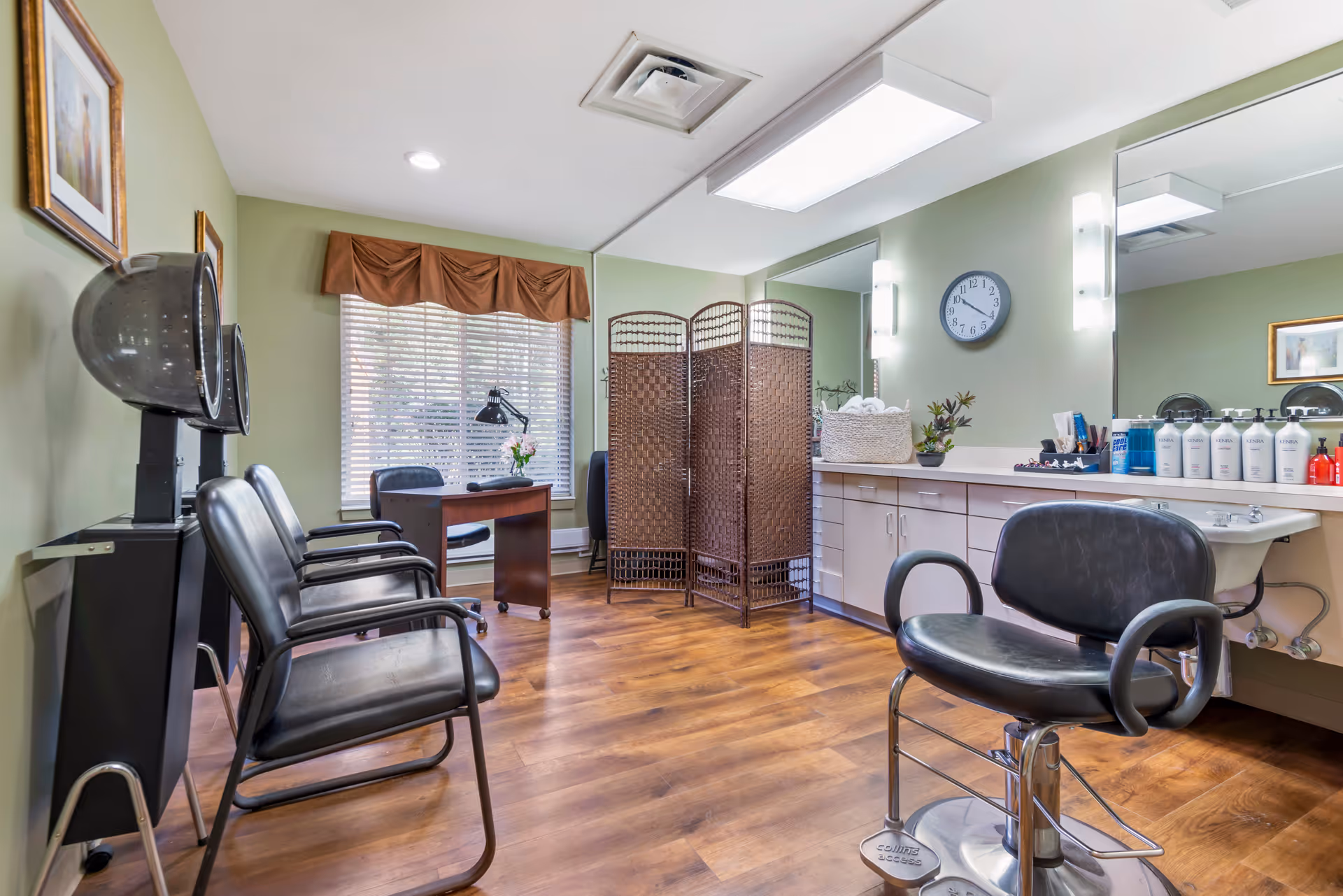 Interior of a hair salon room with green walls and wooden flooring. The room features black salon chairs, hair dryers, a wooden desk with a lamp, a folding privacy screen, a large mirror above a counter with various hair care products, and a wall clock showing 9:10.