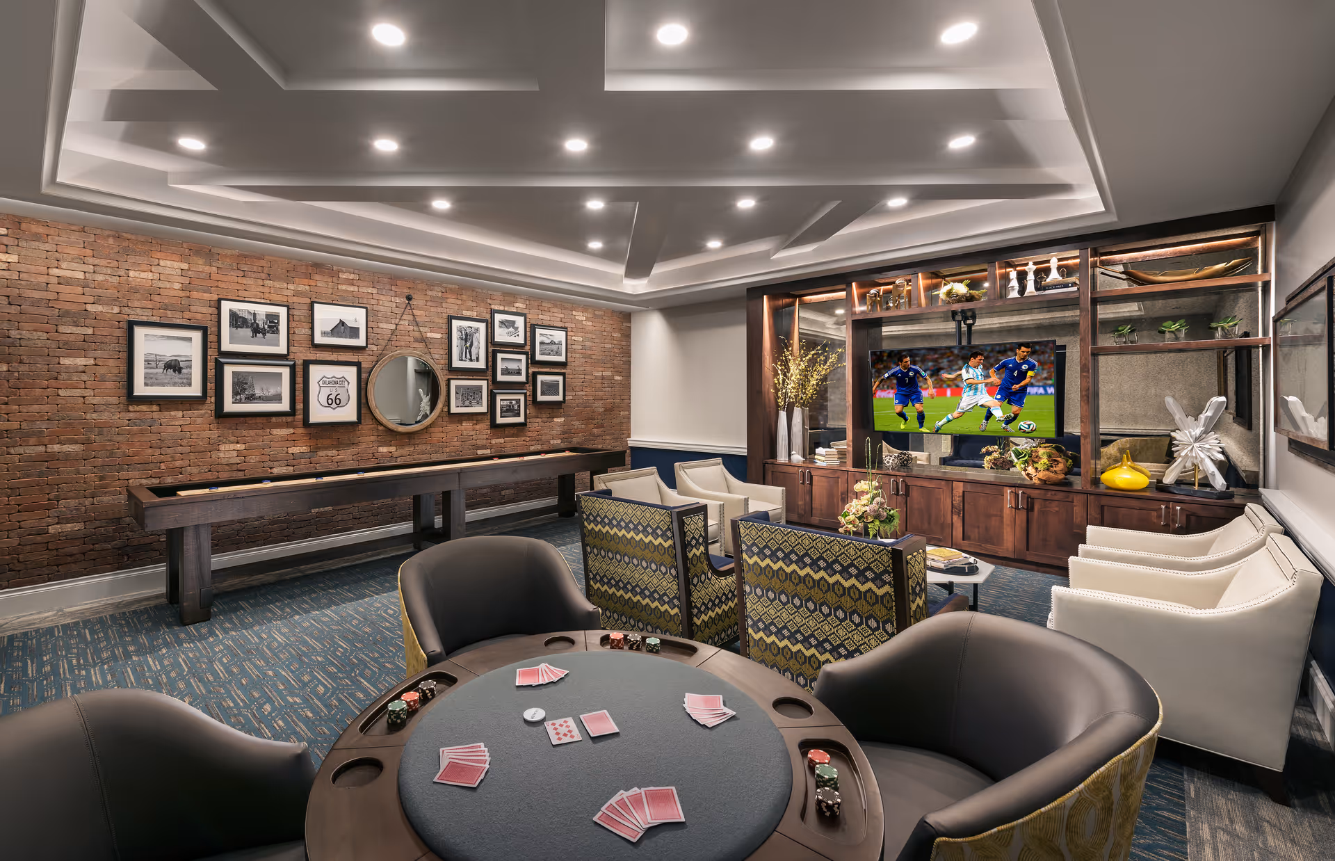 Comfortable lounge and game room with a poker table in the foreground, shuffleboard along a brick wall, and a TV in a built-in entertainment center.