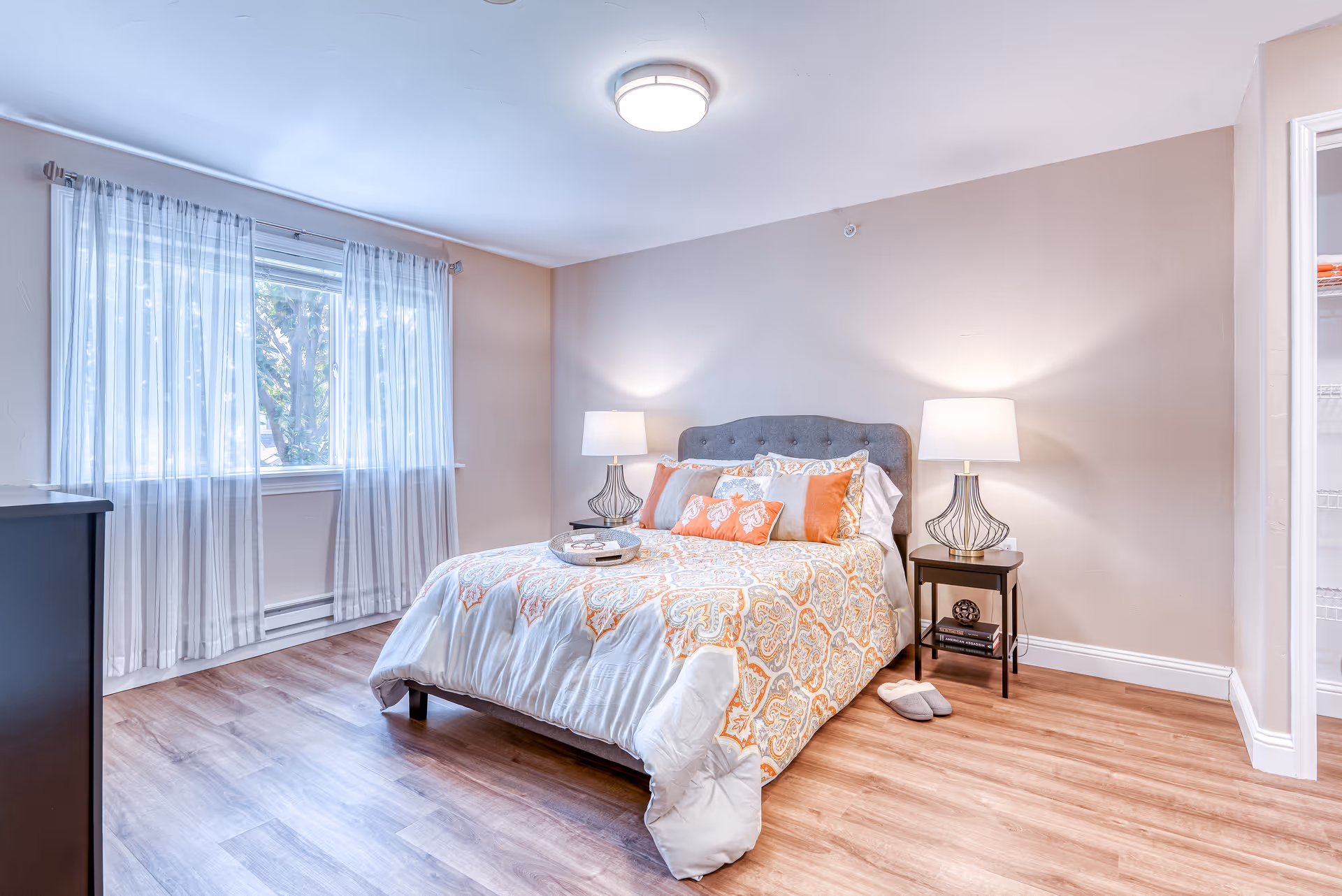 A bright and cozy bedroom with a large window covered by sheer curtains. The room features a bed with a patterned orange and white comforter, multiple pillows, and a tufted gray headboard. There are two bedside tables, each with a lamp, and a pair of slippers on the wooden floor next to the bed. An open closet is visible on the right side of the image.