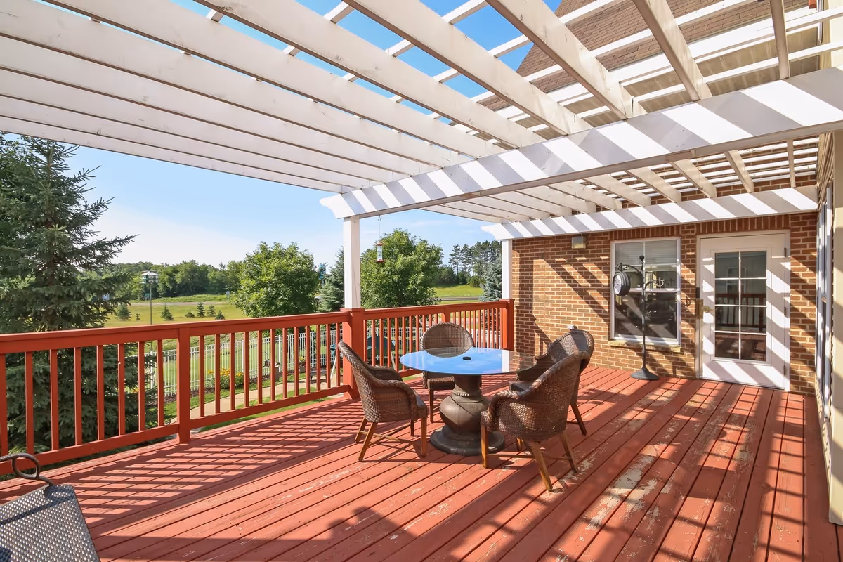 Outdoor patio area with a red wooden deck, a white pergola overhead casting striped shadows, a round glass table with four wicker chairs, and a brick wall with a window and a door leading inside. Green trees and a grassy area are visible in the background under a clear blue sky.