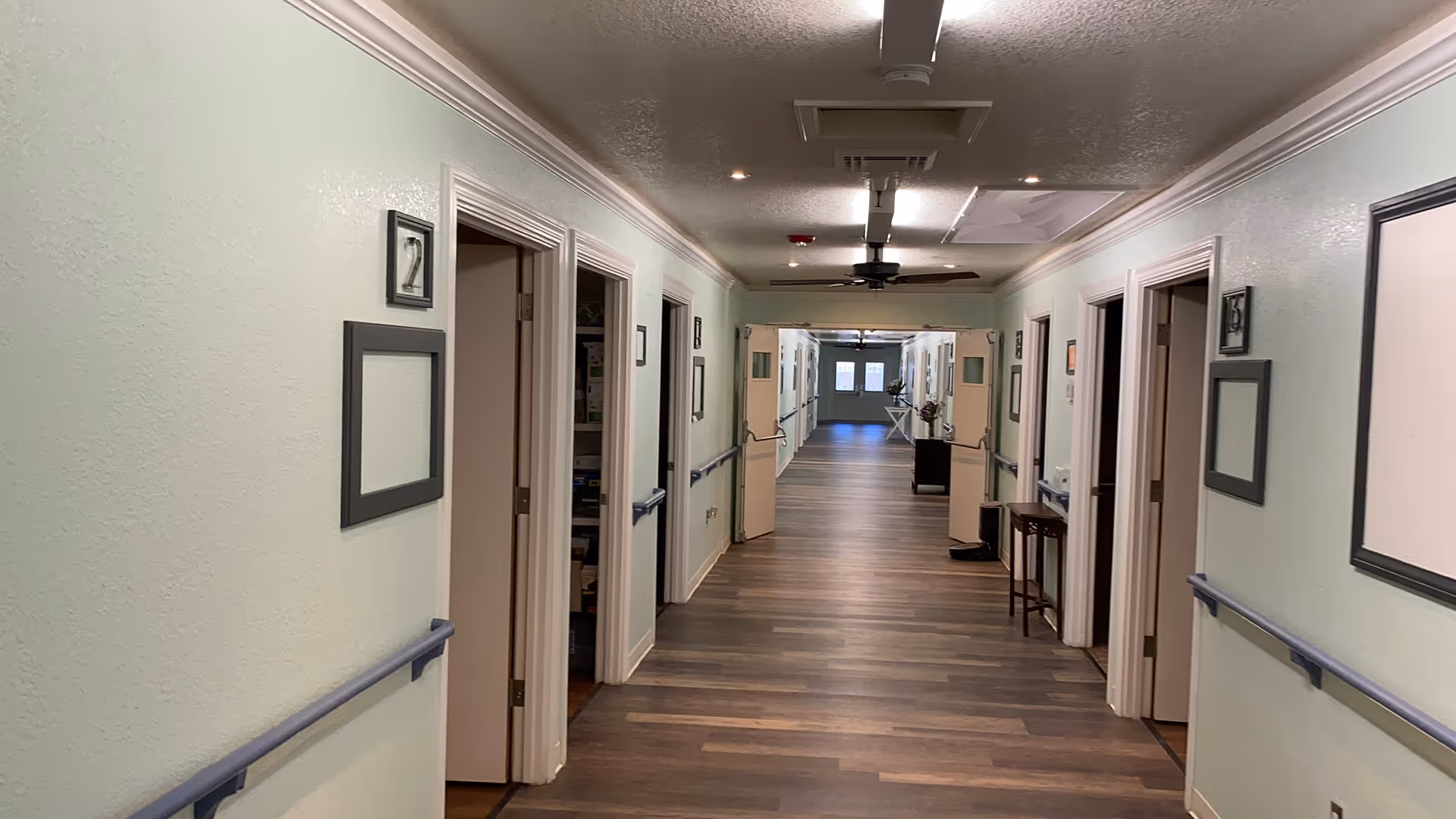 A long, well-lit hallway in a senior living facility with light green walls, wooden flooring, and several open doorways on both sides. The hallway has handrails along the walls, ceiling fans, and double doors at the far end with windows letting in natural light. There are picture frames on the walls and small tables with flowers near the doors.