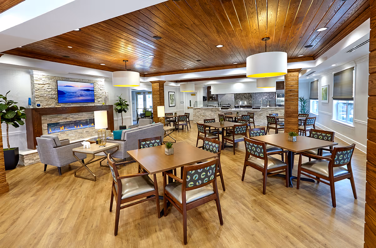 A spacious and well-lit common area in an assisted living facility featuring wooden floors and ceiling with recessed lighting. The room includes several wooden tables with chairs, a seating area with gray sofas and side tables, a modern fireplace with a TV mounted above it, and a kitchen area in the background. Large windows with blinds allow natural light to enter the space.