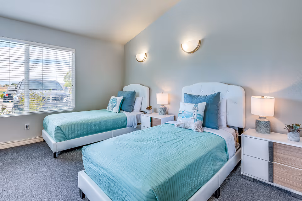 Bright twin bedroom with two white upholstered beds dressed in teal bedding, bedside tables and lamps, and a large window.