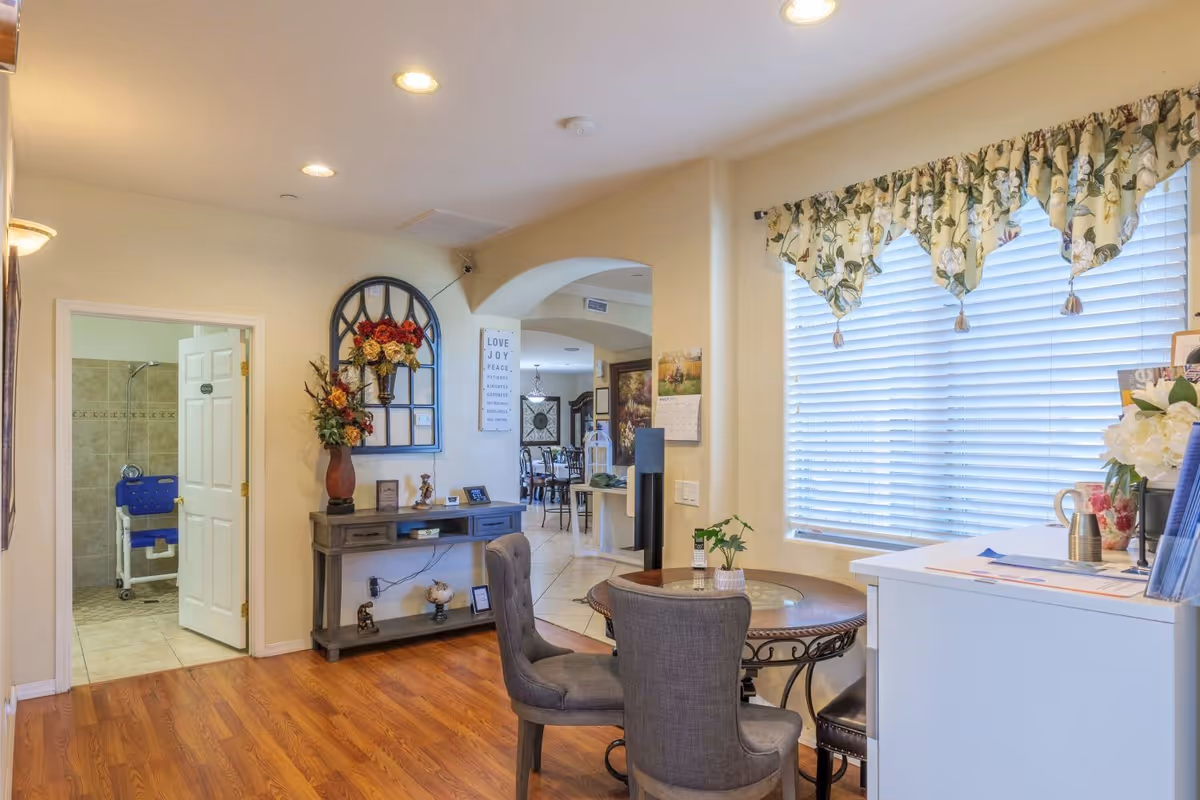 Bright common living area with a round table and chairs by a large window, a decorative console table against the wall, and an open doorway showing a bathroom with a shower chair.