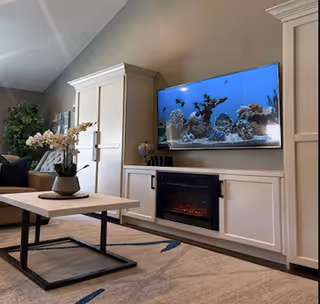 A cozy living room with a mounted flat-screen TV displaying an underwater scene with coral and fish. Below the TV is a white built-in cabinet with an electric fireplace. A modern coffee table with a potted orchid sits on a patterned rug. A beige sofa and a green plant are visible in the background.