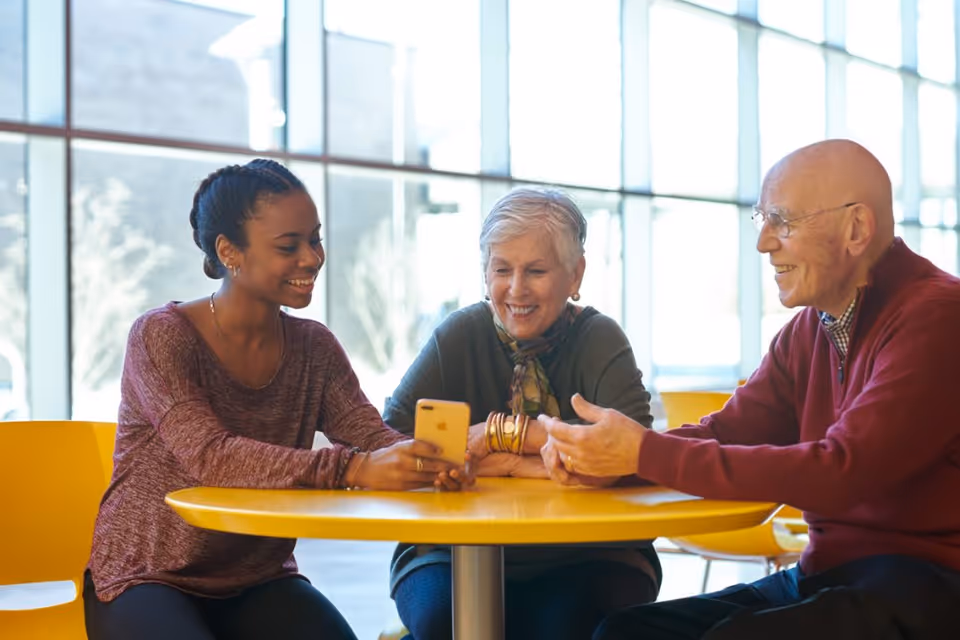 A young woman and two older adults sit at a yellow table in a bright common area, smiling as they look at a smartphone.