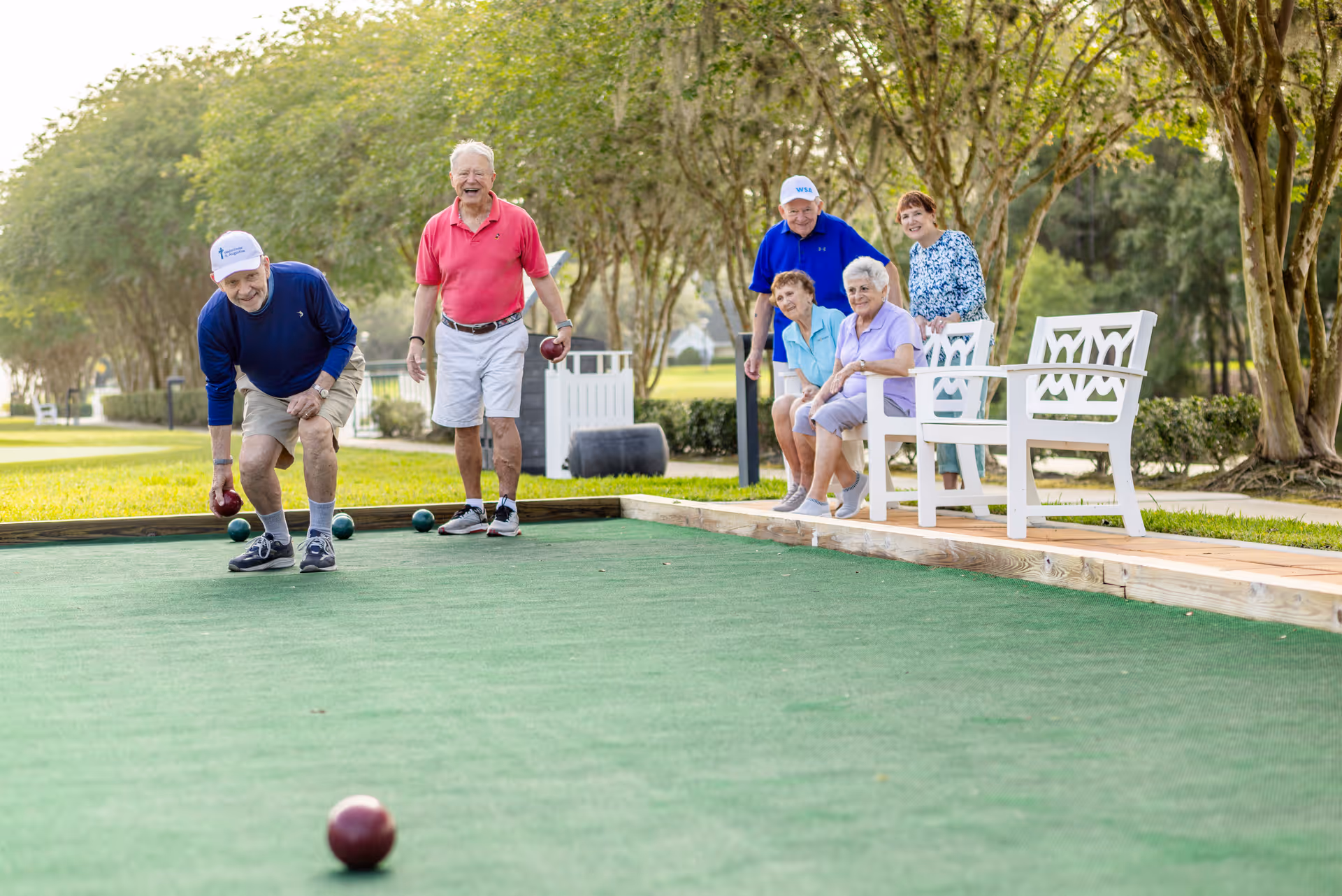 A group of six elderly people enjoying a game of bocce ball outdoors on a green court. Two men are actively playing while four others, including two women sitting on white benches and two people standing, watch and smile. The setting is a sunny day with trees and grass in the background.