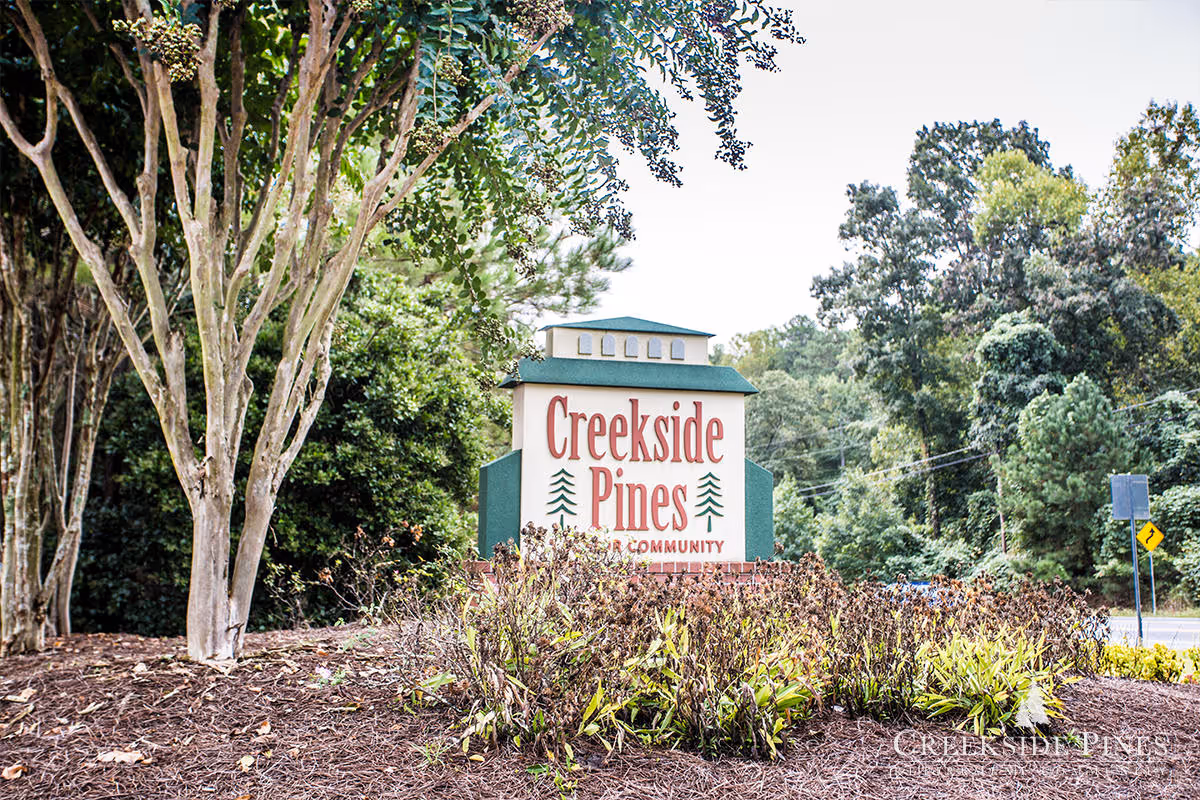 Outdoor view of the Creekside Pines Retirement Community entrance sign surrounded by trees and landscaping with a road visible in the background.