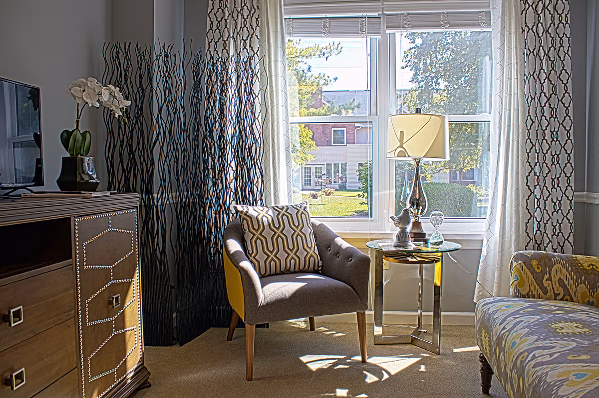 A cozy living room area with a gray armchair featuring a patterned cushion, a glass side table with a decorative lamp and figurines, a patterned sofa, and a wooden cabinet with a TV and a potted orchid. Large windows with patterned curtains allow natural light to brighten the room and provide a view of a green outdoor space with trees and buildings.