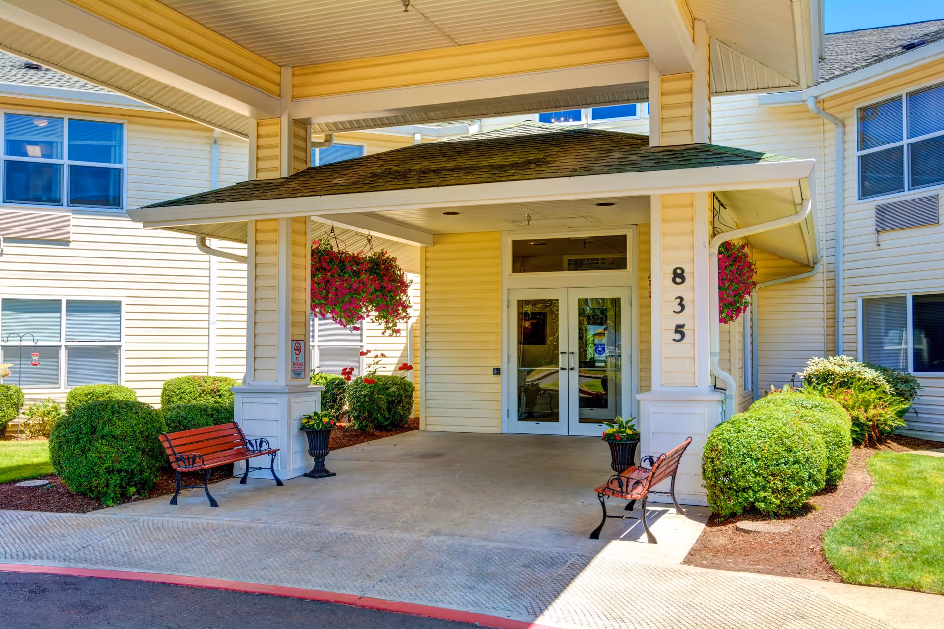Entrance to a senior living facility with a covered drop-off area, two benches, hanging flower baskets, and well-maintained bushes and lawn. The building is light yellow with white trim and has the number 835 displayed near the door.