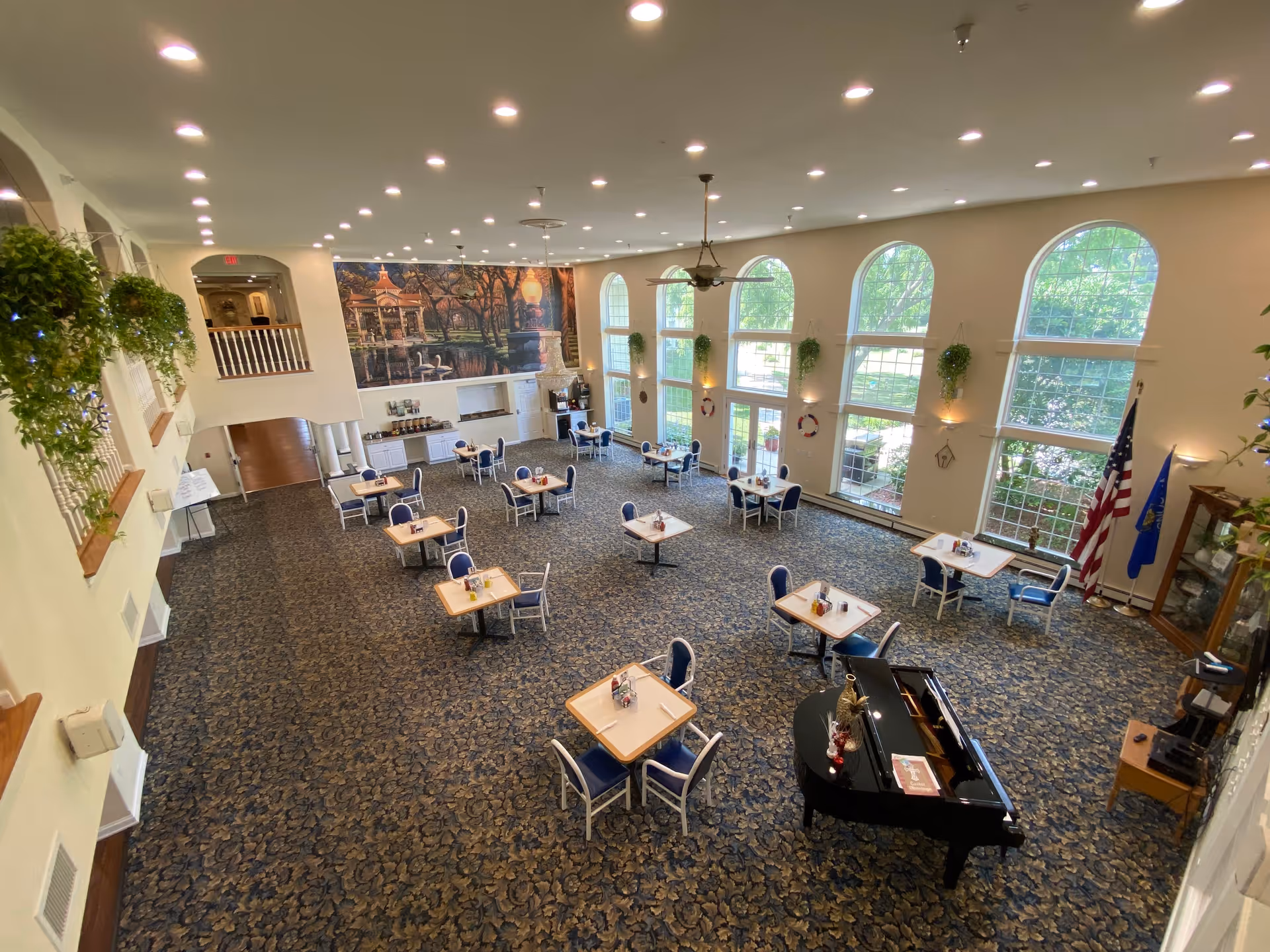 Spacious dining area with multiple small square tables each surrounded by four blue cushioned chairs. Large arched windows line one wall, allowing natural light to fill the room. A black grand piano is positioned near the windows. The room has a patterned carpet, hanging plants, and a large mural on the far wall depicting a gazebo in a park setting. Flags and a display cabinet are visible near the windows.