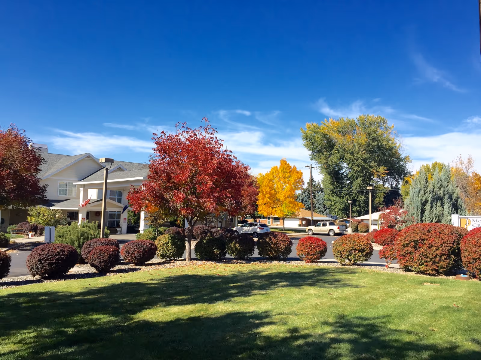 Exterior view of McKay Creek Assisted Living facility with a well-maintained lawn, various bushes and trees with autumn foliage, a clear blue sky, and a parking area with several cars.