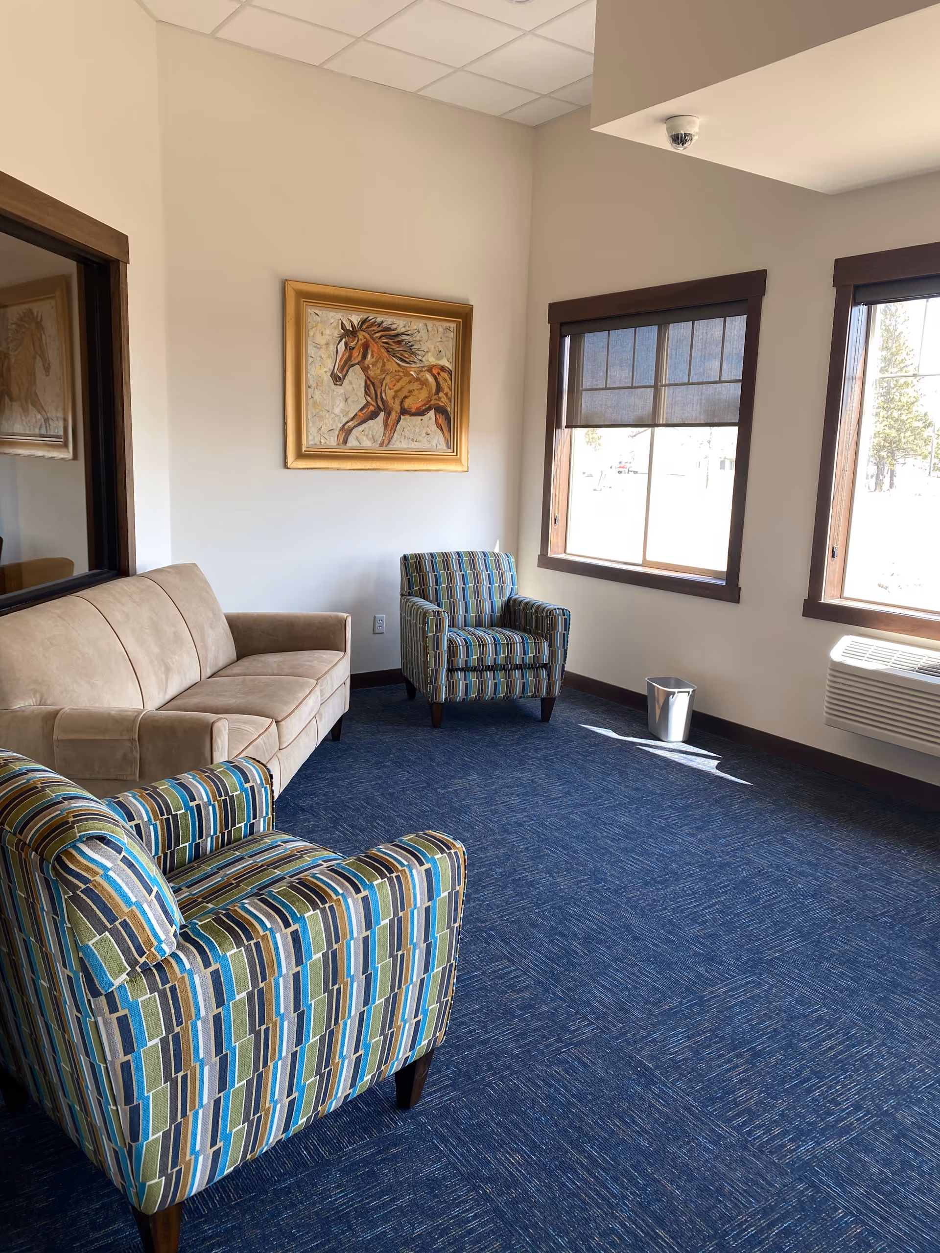 A small sitting area with a beige sofa and two patterned armchairs on a blue carpet. There are two windows with dark brown frames and blinds, a wall-mounted air conditioning unit below one window, a small silver trash can on the floor, and a framed painting of a horse on the wall.
