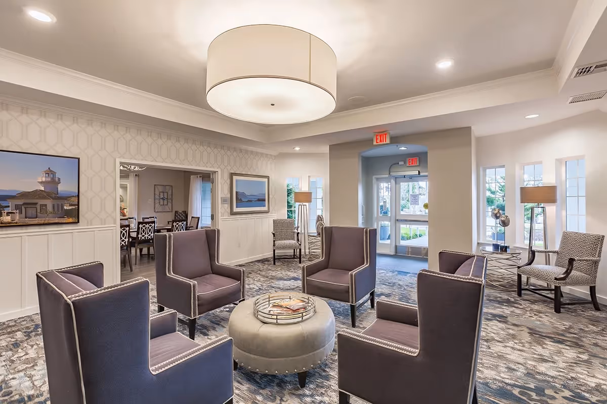 A well-lit senior living facility common area with four purple armchairs arranged around a round ottoman. The room has patterned carpet, a large ceiling light fixture, and framed pictures on the walls. There is a doorway leading to a dining area with tables and chairs, and an exit door with windows letting in natural light.