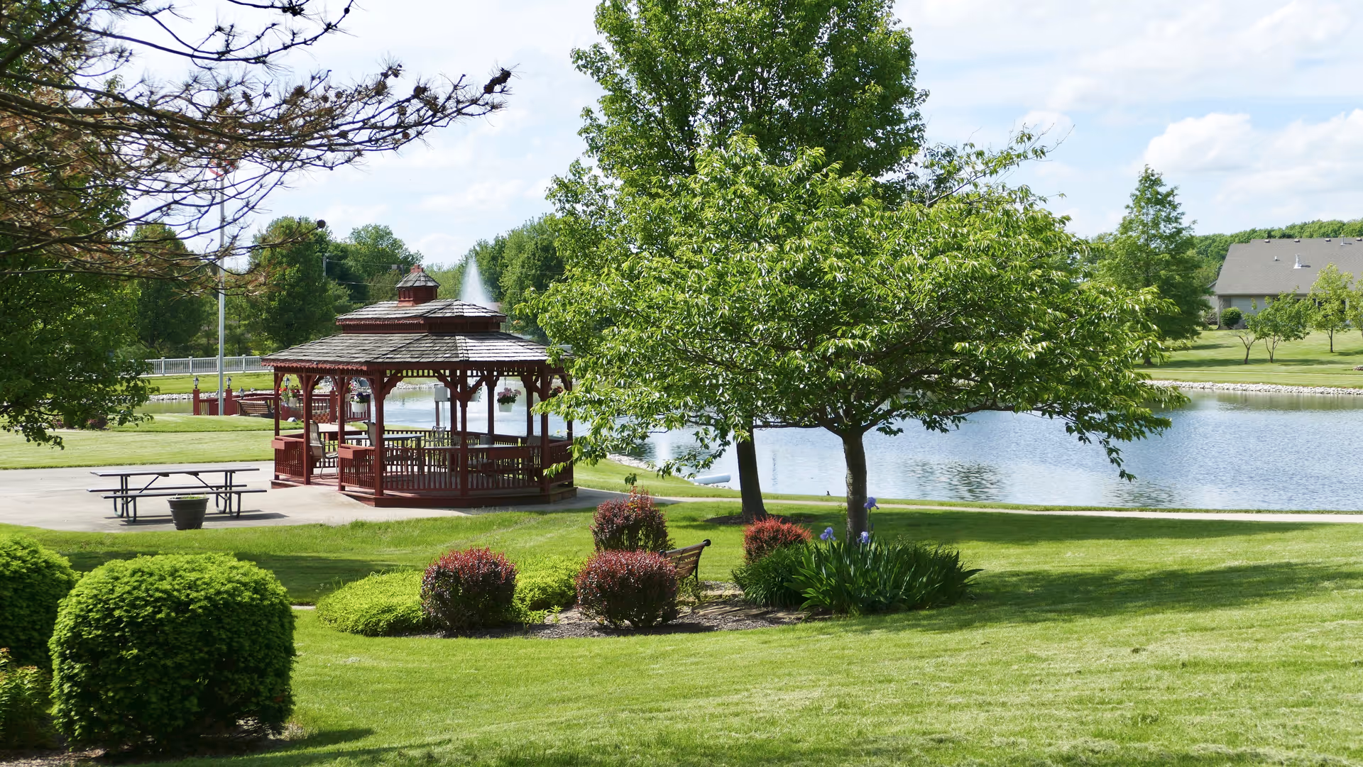 A peaceful outdoor scene at The Cedars featuring a red wooden gazebo near a pond with a fountain. The area is surrounded by green grass, trees, bushes, and a picnic table on a paved surface. A bench is placed near some flowering plants under a tree.