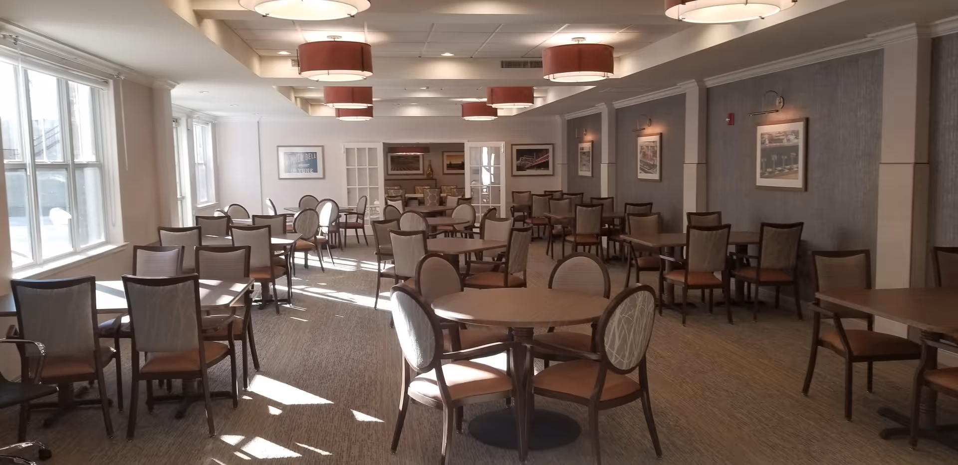 A spacious dining room with multiple round and square tables surrounded by chairs. The room has large windows on one side allowing natural light to fill the space. The walls are decorated with framed pictures, and several ceiling lights with red lampshades hang from the ceiling.