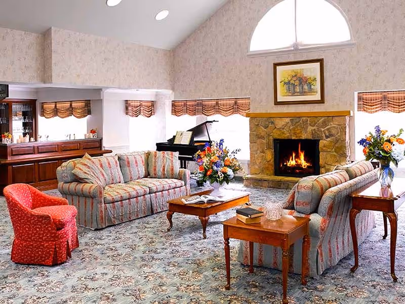 A cozy living room with two striped sofas, a red armchair, a wooden coffee table with books and flowers, a stone fireplace with a fire burning, a grand piano in the corner, and large windows with valances letting in natural light.