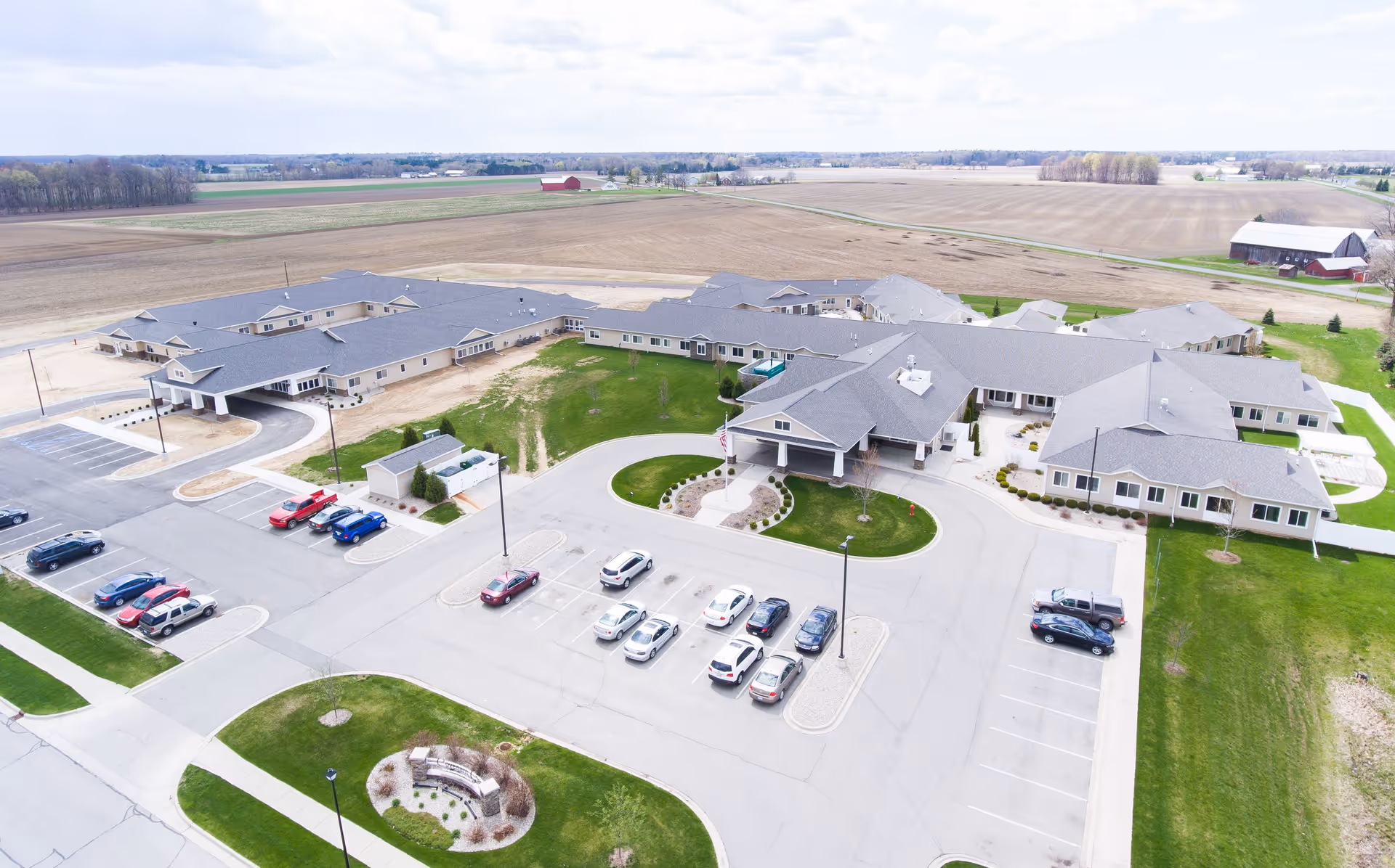 Aerial view of Covenant Glen of Frankenmuth senior living facility showing a large single-story building with multiple wings, surrounded by parking lots with cars, green lawns, and open farmland in the background under a cloudy sky.