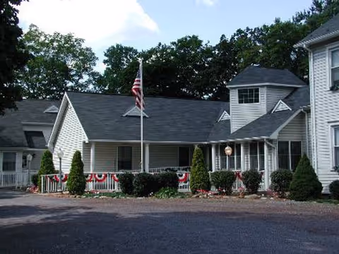 Exterior view of a white multi-section building with a dark roof, surrounded by trees. The building has a porch decorated with red, white, and blue bunting and an American flag on a flagpole in front. There are shrubs and small trees planted along the front of the building.