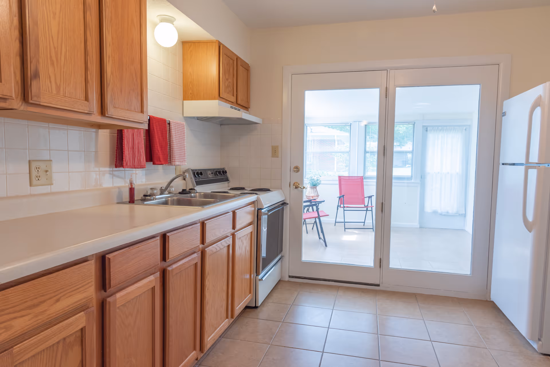A clean kitchen with wooden cabinets, a white countertop, a double sink, an electric stove with an overhead vent, and a white refrigerator. Two red towels hang on the wall above the sink. Glass double doors lead to a bright sunroom with two red chairs and a small table.
